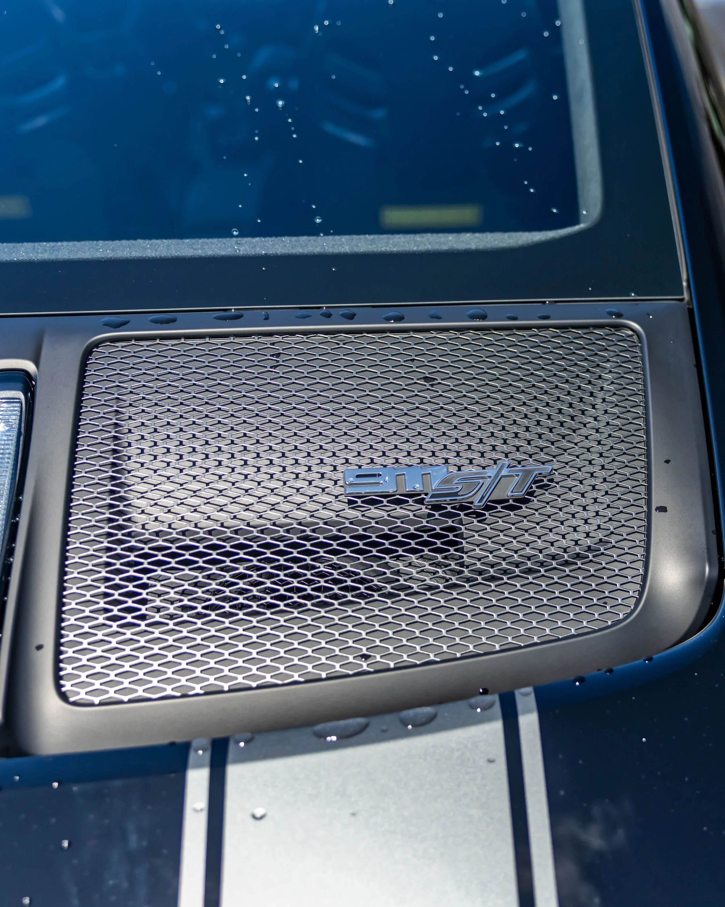 Air | Water 2025 — Orange County Fairgrounds — A high-angle, close-up photograph of a Porsche 911 S/T grill featuring silver racing stripes on the deck lid. The focus is on a black deck lid with a silver mesh grille, which has a chrome '911 S/T' embl