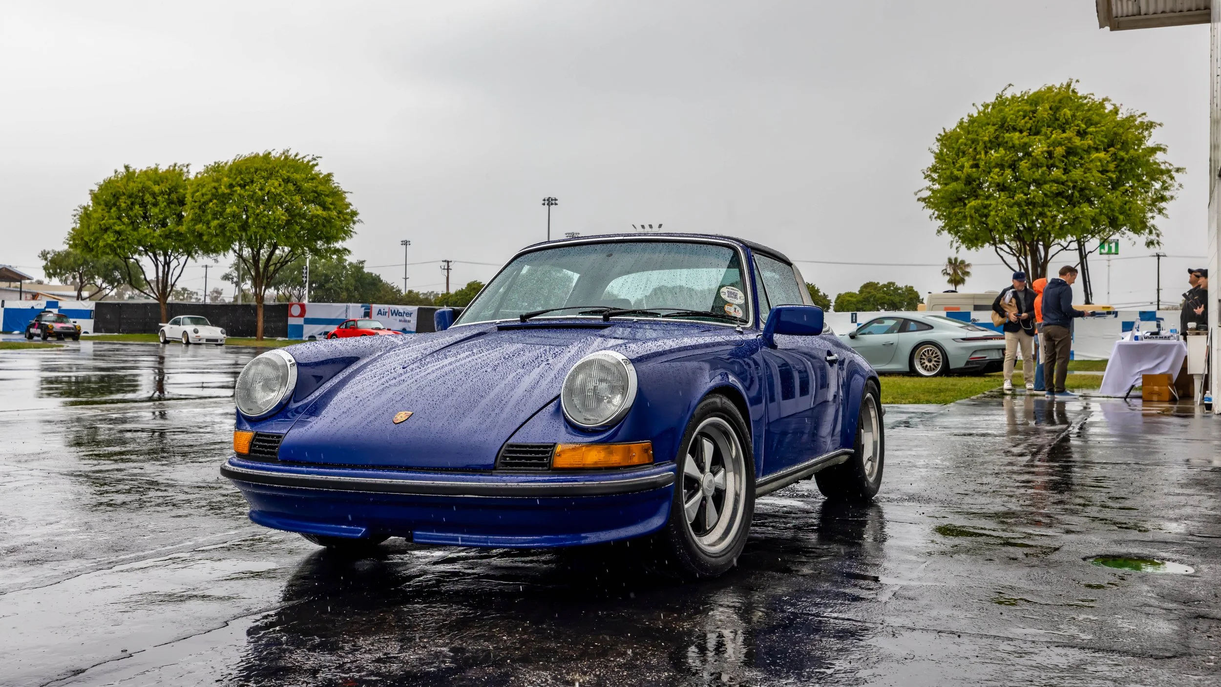 Air | Water 2025 — Orange County Fairgrounds — A classic G-body Porsche 911 Targa in a vibrant blue is parked on wet pavement, covered in raindrops. The photo is taken from a low front three-quarter angle during an overcast day at the 'Air | Water' P