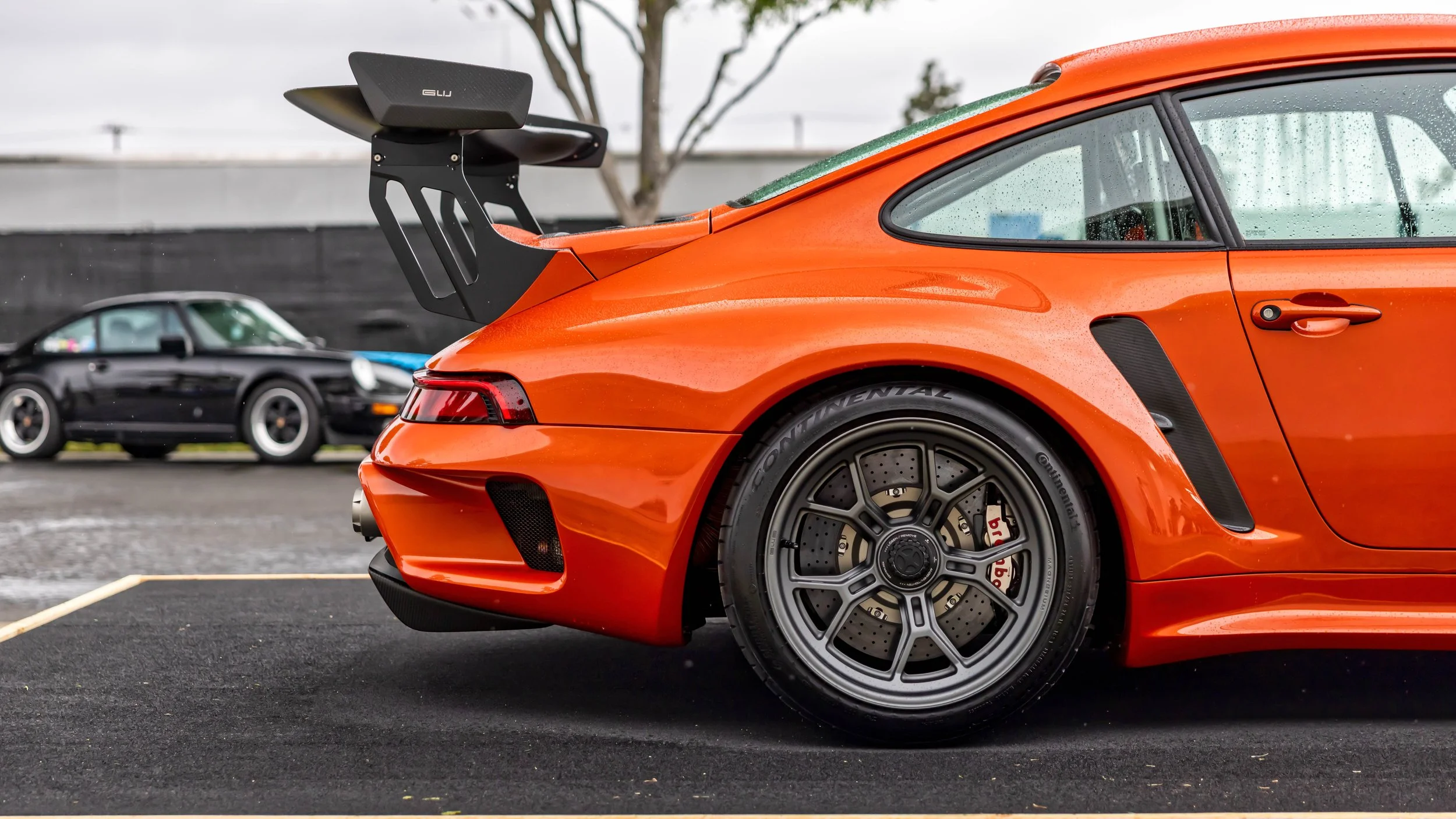 Air | Water 2025 — Gunther Werks — Orange County Fairgrounds — A rear three-quarter view of a Lava Orange Gunther Werks Turbo (Tornado) remastered Porsche 911on wet asphalt on an overcast day. The car is fitted with grey magnesium center-lock wheels,