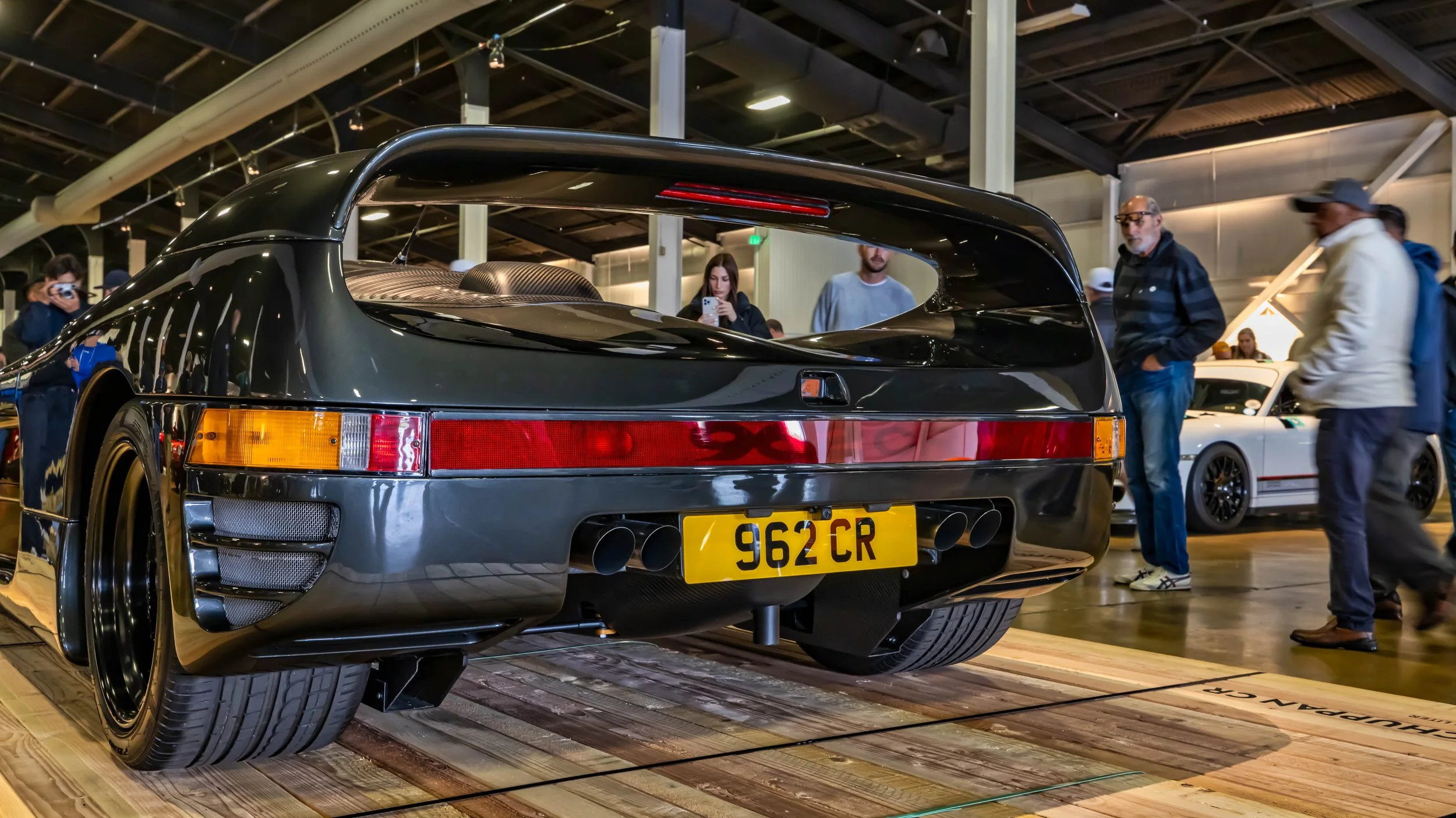 Air | Water 2025 — Orange County Fairgrounds — A low-angle, rear three-quarter view of a dark gray Schuppan 962CR supercar on display at an indoor automotive event. The car, based on the Porsche 962 race car, features a prominent integrated rear wing