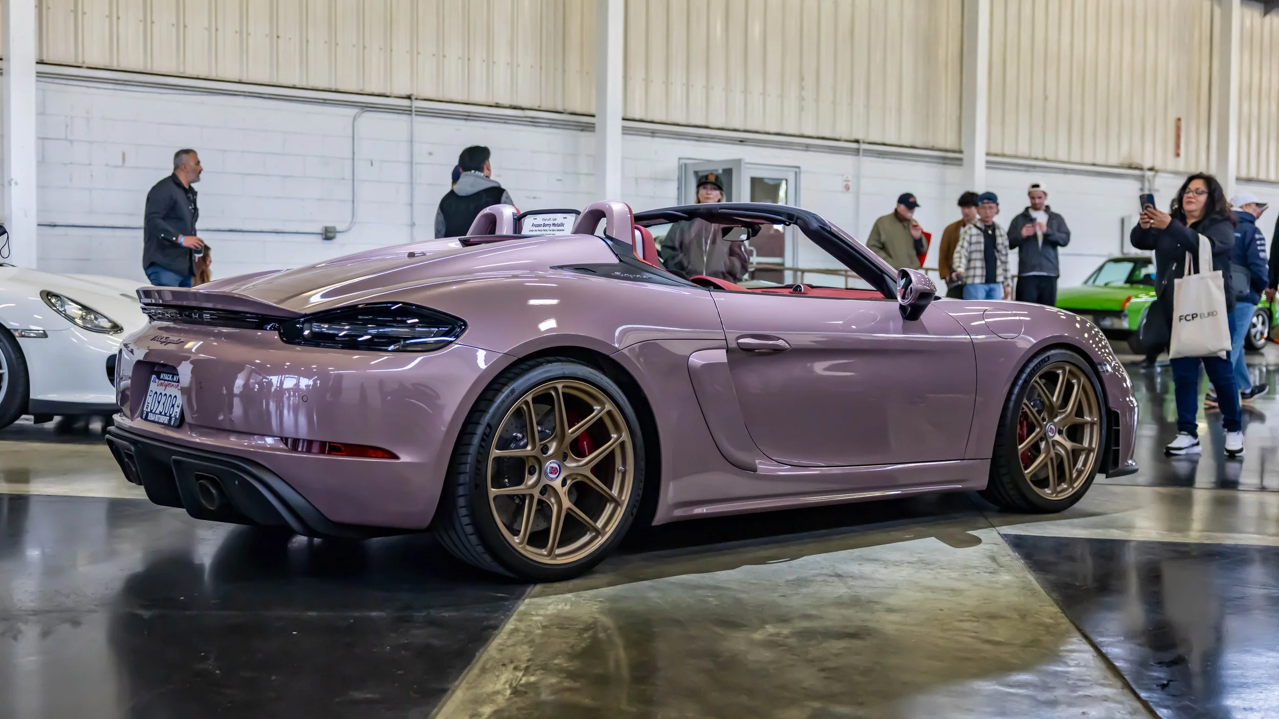 Air | Water 2025 — Orange County Fairgrounds — Matt Farah's Porsche 718 Boxster Spyder in Frozen Berry Metallic is parked on a reflective concrete floor at an indoor car show. The vehicle is shown from the rear three-quarter view, highlighting its cu