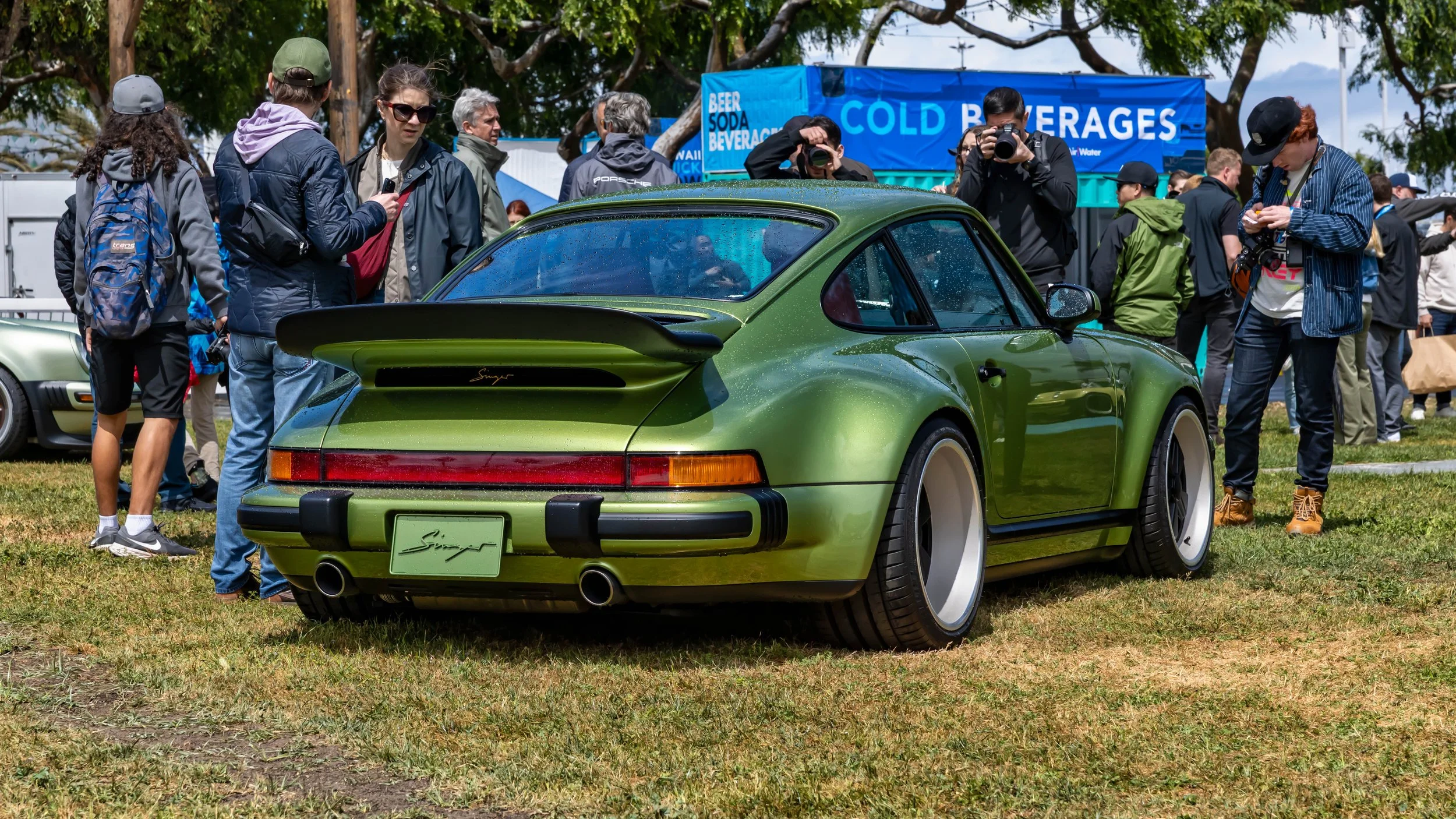 Air | Water 2025 — Orange County Fairgrounds — A metallic linden green Porsche 911 Reimagined by Singer is parked on a grassy field, viewed from the rear three-quarter angle. The car features a prominent 'whale tail' spoiler, wide rear fenders, and d
