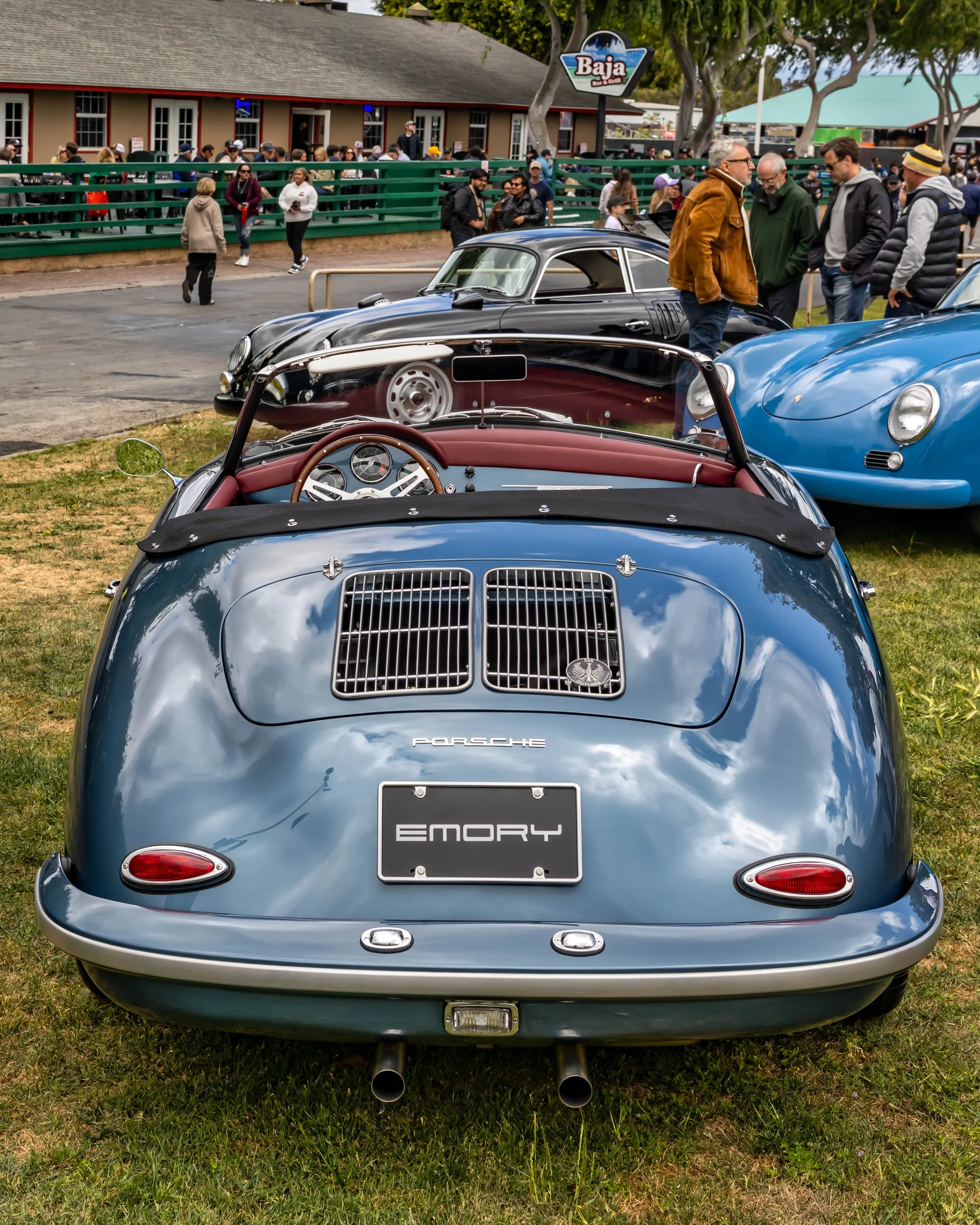 Air | Water 2025 — Emory Motorsports — OC Fairgrounds — A rear view of a blue-grey Emory Outlaw Porsche 356 Speedster parked on a grassy lawn at an outdoor car event. The car features a red leather interior, a wood-rimmed steering wheel, and a custom