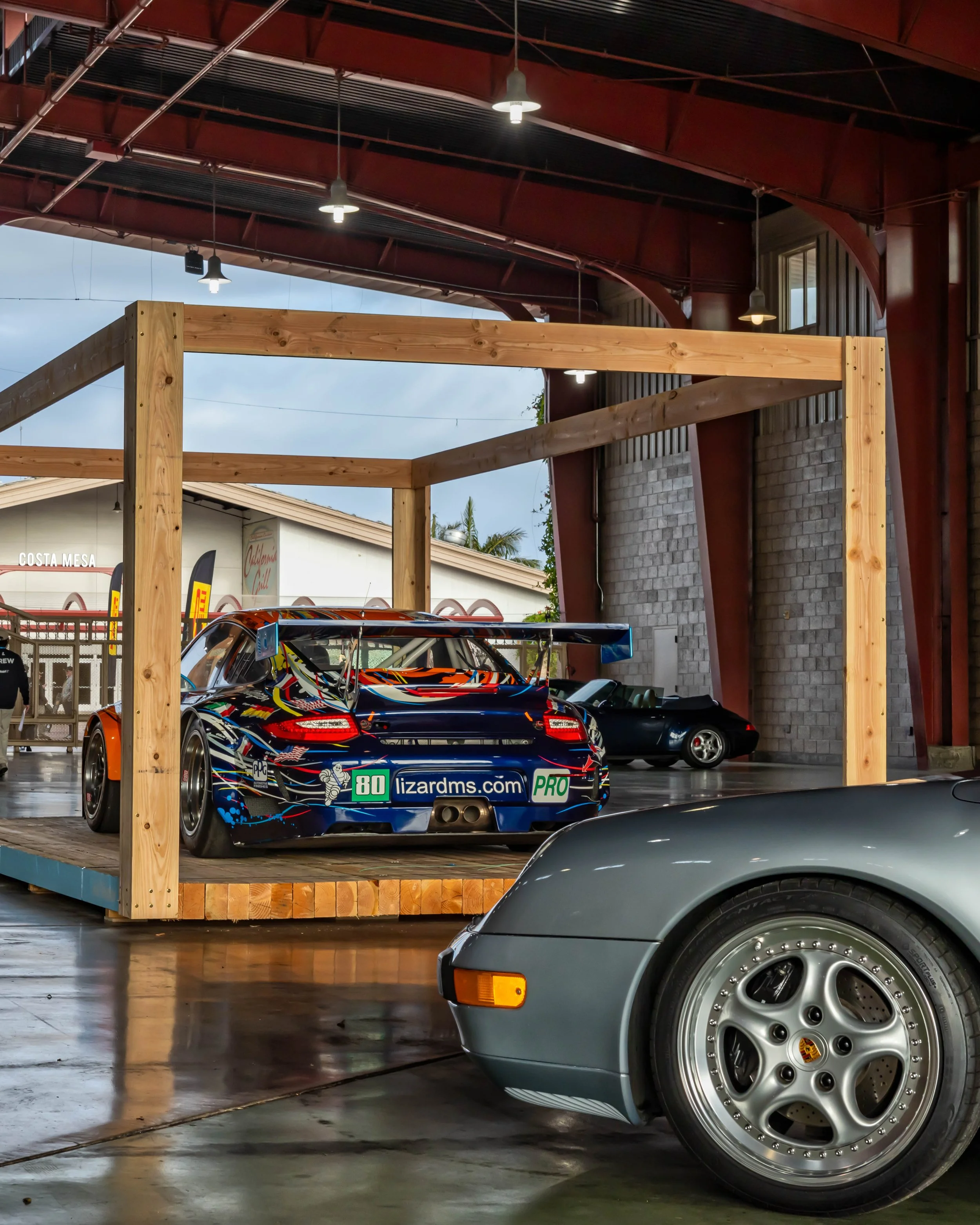 Air | Water 2025 — Orange County Fairgrounds — A low-angle shot inside an exhibition event space, focusing on a Porsche 997 GT3 RSR race car with a vibrant 'art car' livery, displayed on a wooden platform. In the foreground, the front fender and mult