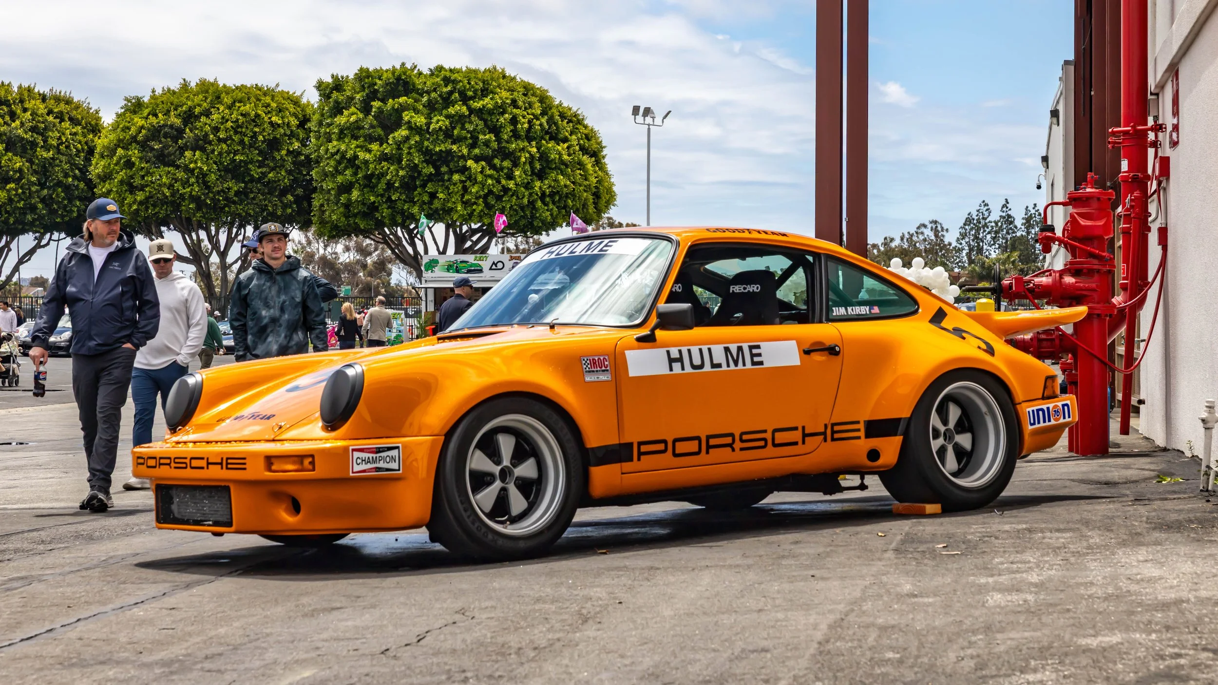 Air | Water 2025 — Orange County Fairgrounds — A low-angle, front three-quarter view of a 1974 Porsche 911 RSR IROC tribute car, finished in bright orange with a full racing livery. The car, featuring a prominent 'whale tail' spoiler and Fuchs wheels