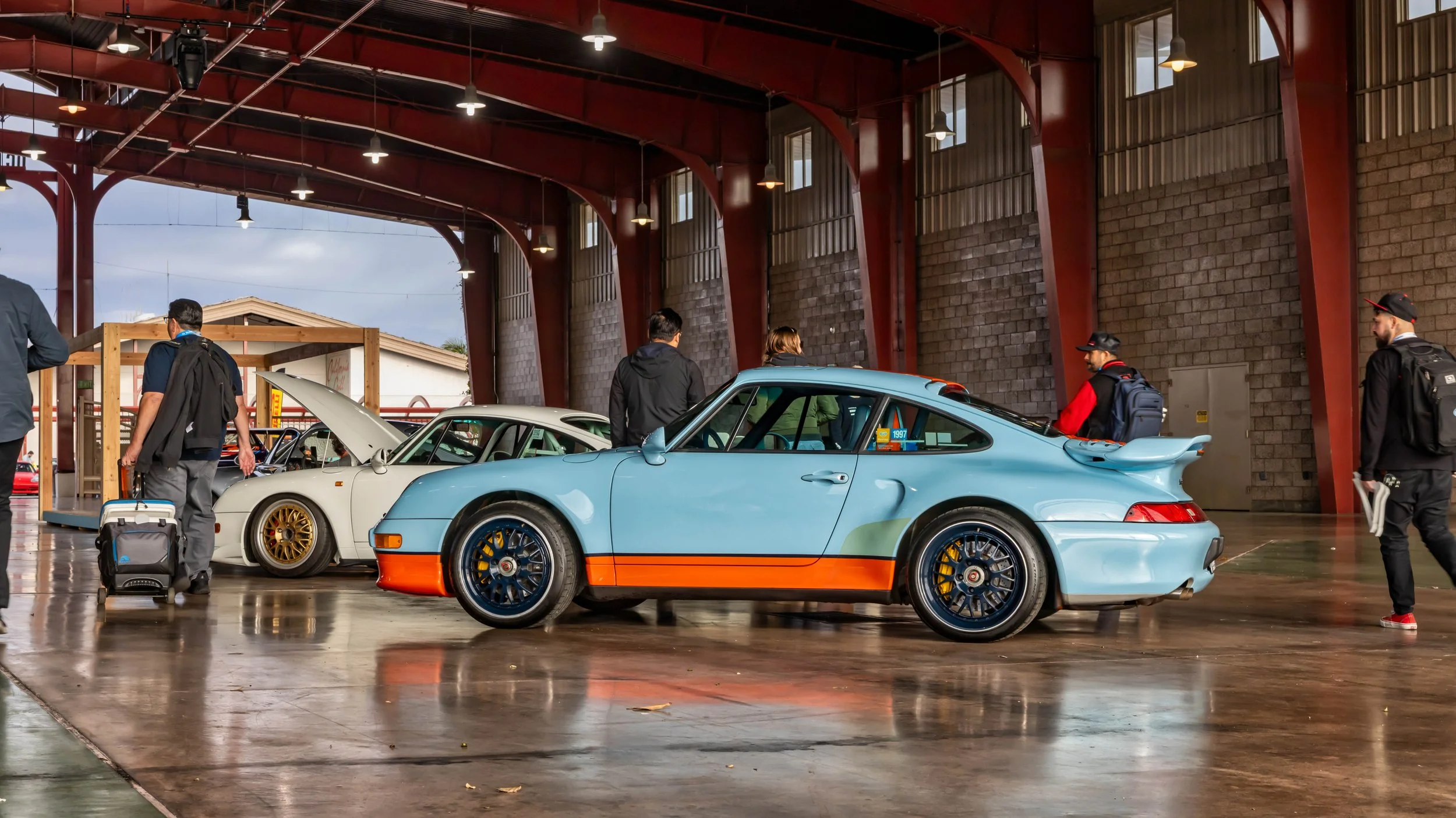 Air | Water 2025 — Orange County Fairgrounds — A Porsche 993 finished in a light blue and orange Gulf-style livery is displayed indoors on a polished concrete floor. The car features dark blue multi-spoke wheels with yellow brake calipers and a large