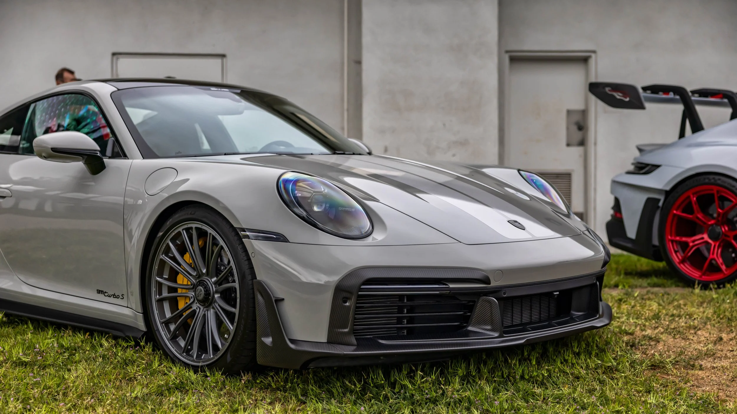 Air | Water 2025 — Gunther Werks — Orange County Fairgrounds — A Chalk Gray Porsche 911 Turbo S (992) is parked on a grassy area, viewed from a low, front three-quarter angle. The car is equipped with a GW09 carbon-fiber front spoiler, dark gray mult