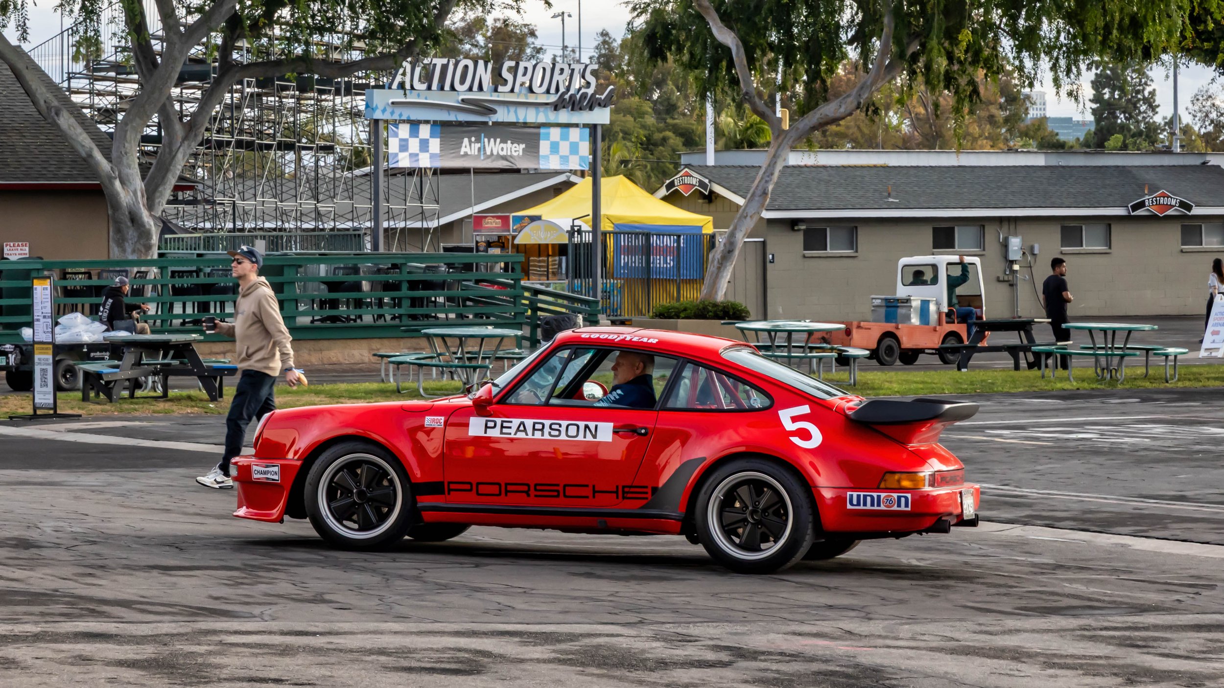 Air | Water 2025 — Orange County Fairgrounds — A red Porsche 911 RSR IROC tribute car, number 5, with a prominent 'whale tail' spoiler, is driving through a paved event area. The car features a racing livery with 'PORSCHE', 'PEARSON', and 'GOODYEAR' 