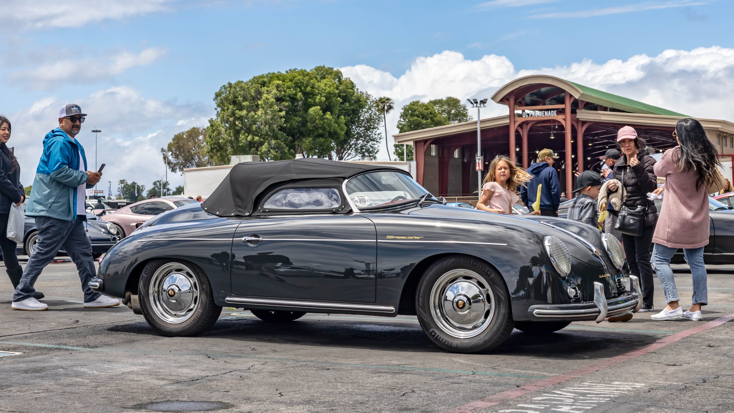 Air | Water 2025 — Orange County Fairgrounds — A dark grey classic Porsche 356 Speedster with its black soft top raised is parked on an asphalt lot during a daytime car event. Spectators are gathered around the vehicle, with a building labeled "OC PR