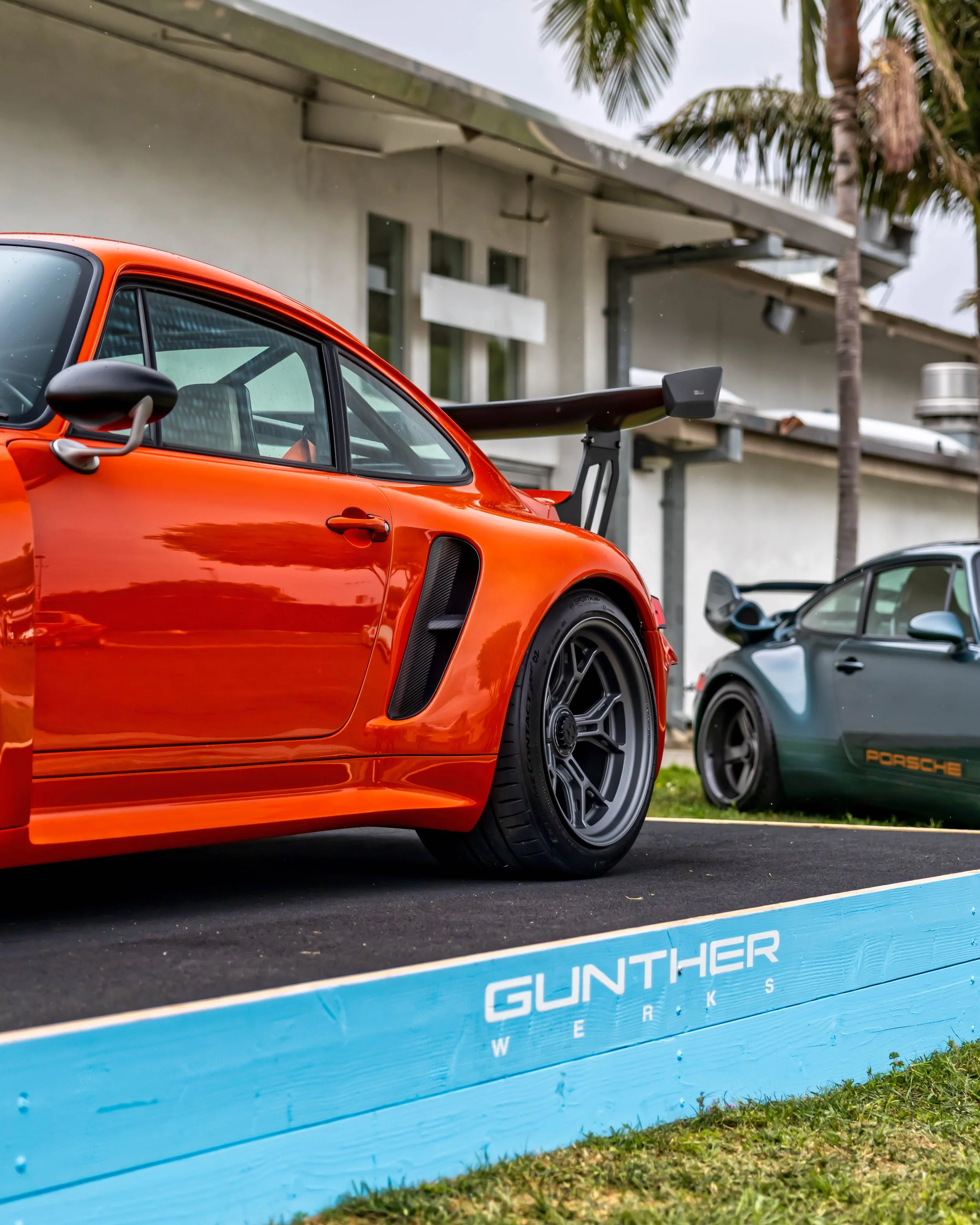 Air | Water 2025 — Gunther Werks — Orange County Fairgrounds — A low-angle, rear three-quarter view of a vibrant orange Gunther Werks Turbo (Tornado), a Remastered Porsche 993 on a blue display platform. The car features a large rear wing, carbon-fib