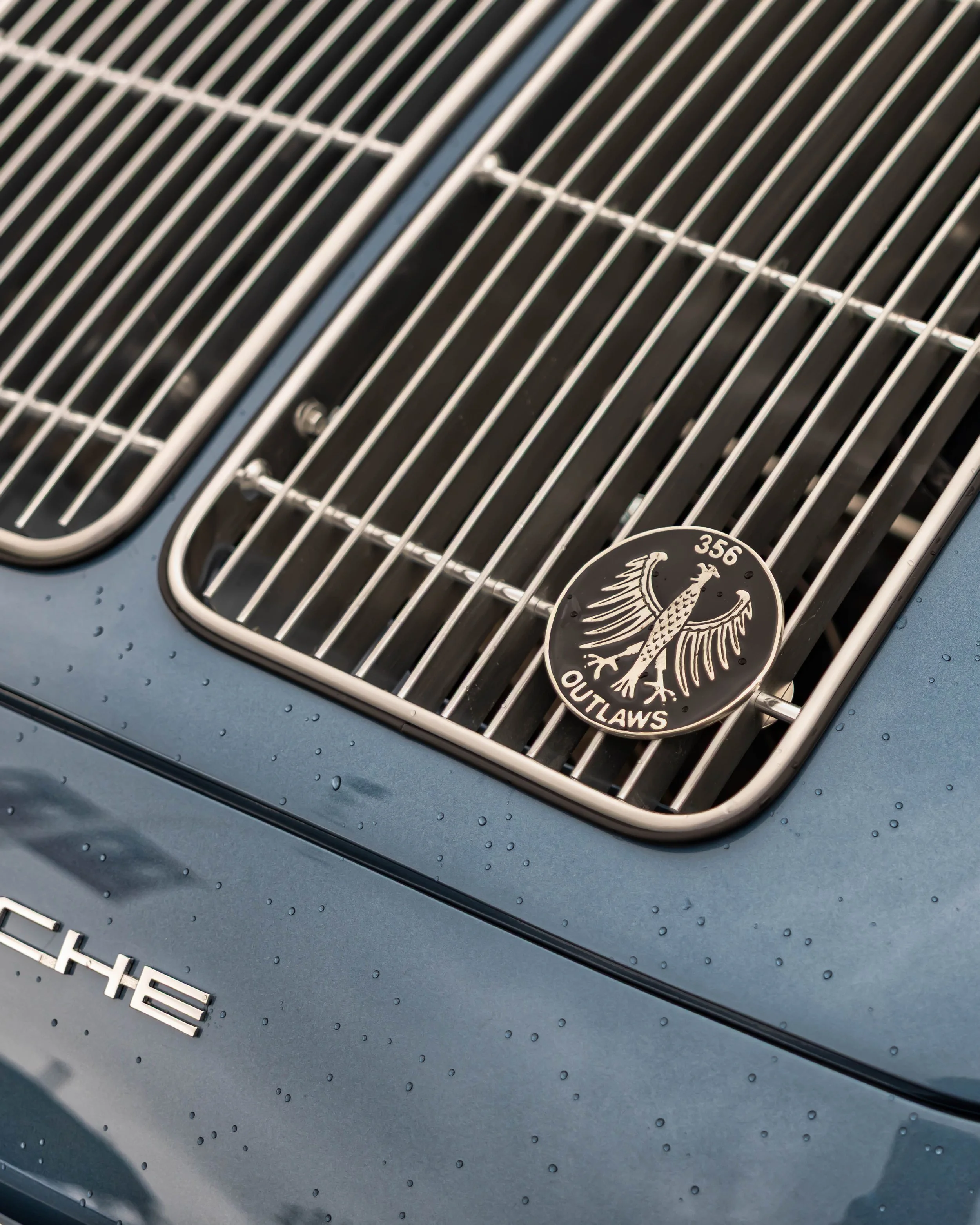 Air | Water 2025 — Emory Motorsports — OC Fairgrounds — A detailed close-up of the rear engine lid of a classic Porsche 356 Emory Outlaw, finished in a metallic blue-gray paint beaded with water droplets. The shot highlights the twin chrome engine gr