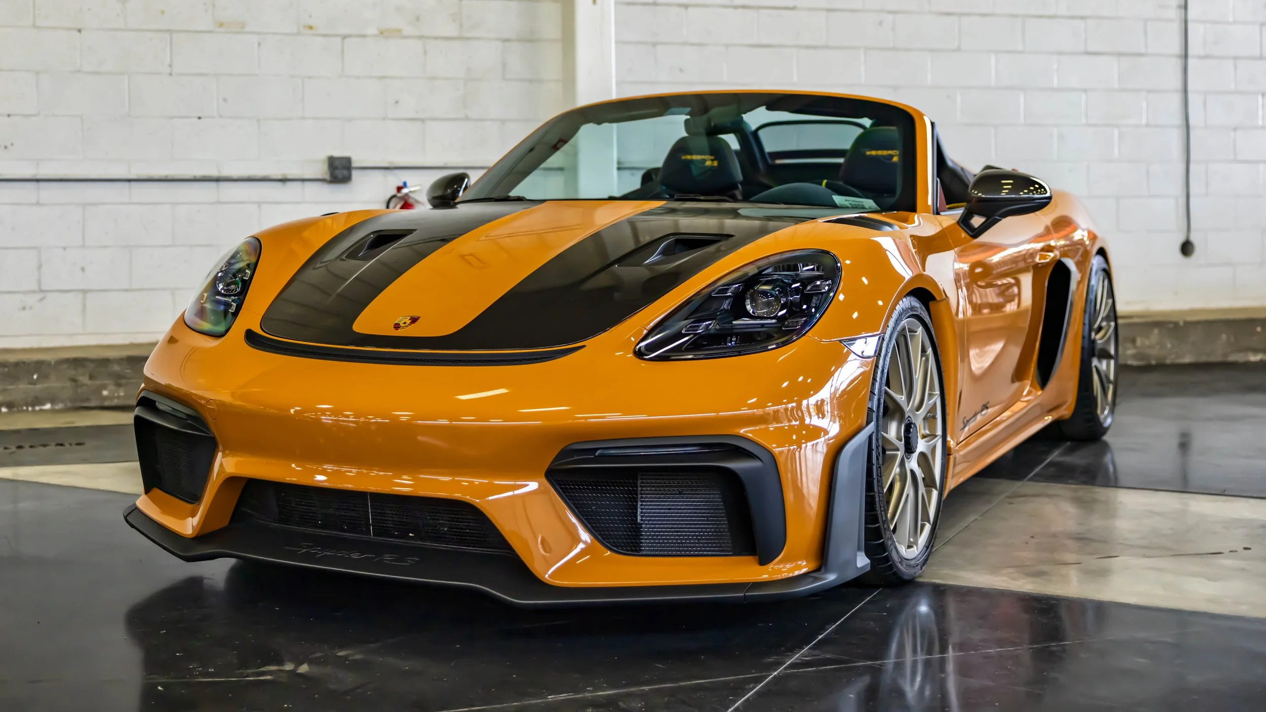 Air | Water 2025 — Orange County Fairgrounds — A 2024 Porsche 718 Spyder RS in a vibrant Signal Yellow finish, featuring a prominent black center stripe over its carbon-fiber hood with dual NACA ducts. The car is parked indoors on a reflective black 