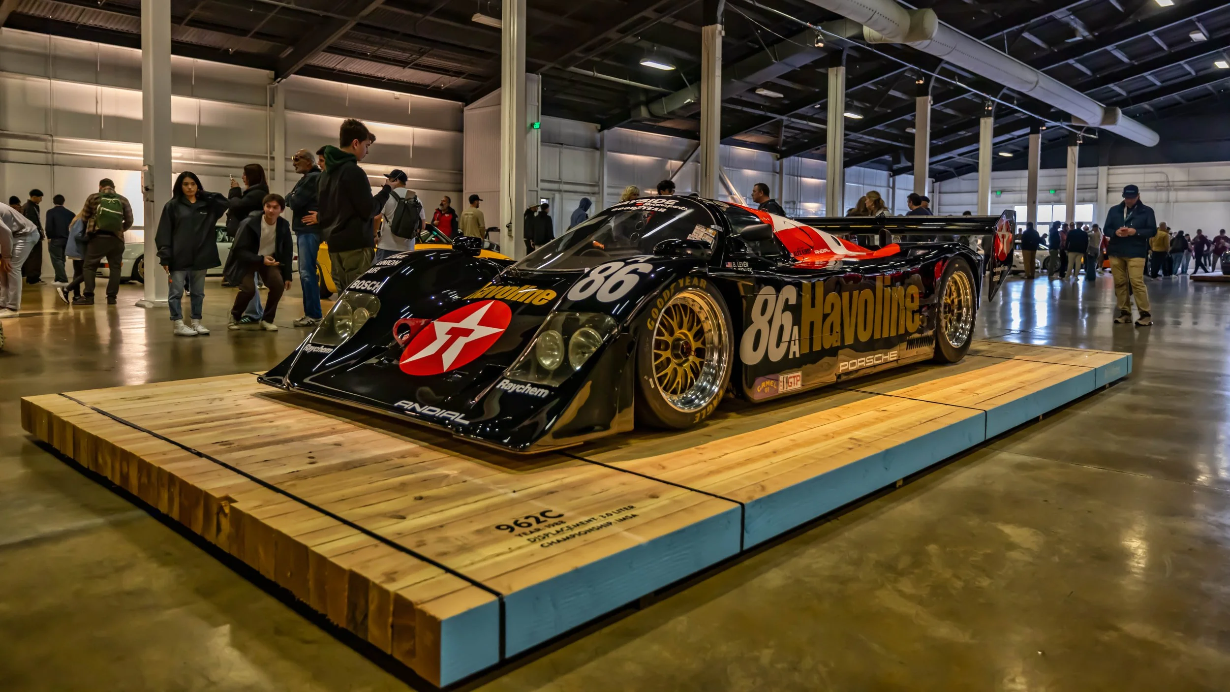Air | Water 2025 — Orange County Fairgrounds — A low-angle, front three-quarter view of a 1988 Porsche 962C race car, number 86, featuring the iconic black, red, and gold Havoline livery. The car, fitted with gold BBS-style multi-spoke racing wheels,