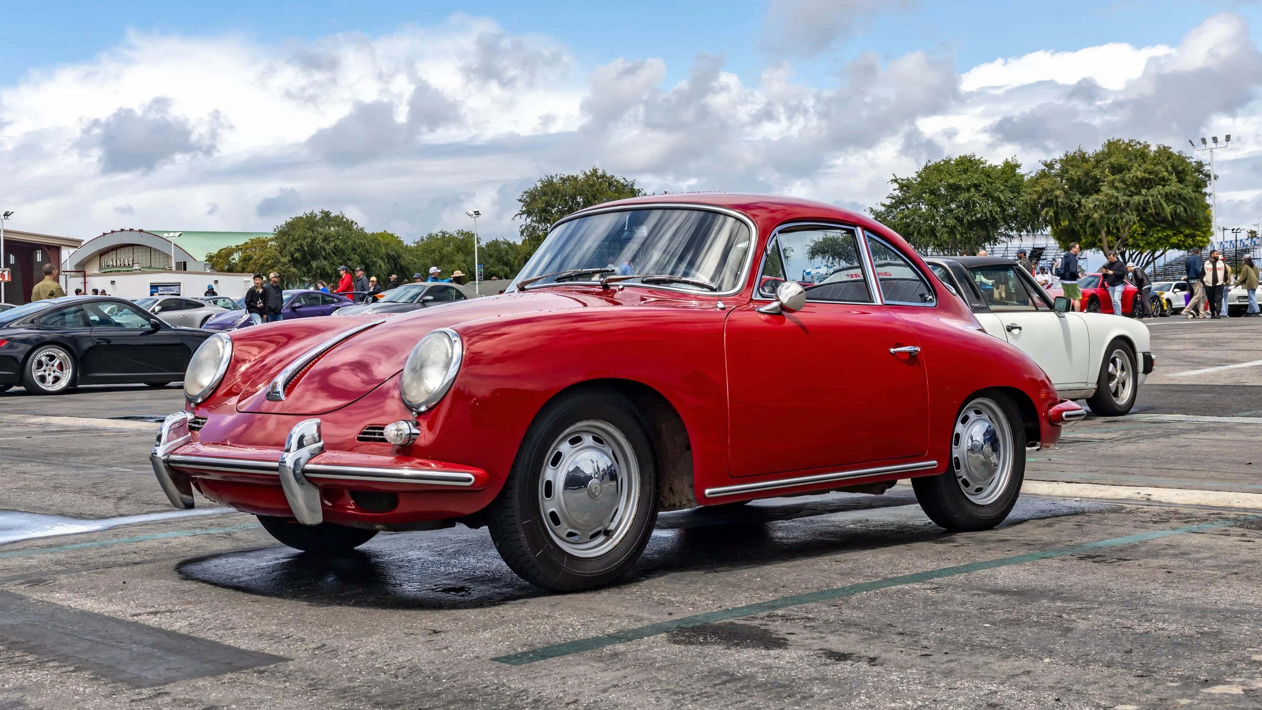 Air | Water 2025 — Orange County Fairgrounds — A classic, vibrant red Porsche 356C Coupe is parked at an outdoor car gathering on a cloudy day. The car is shown from a low, front three-quarter angle, highlighting its iconic rounded shape, chrome bump