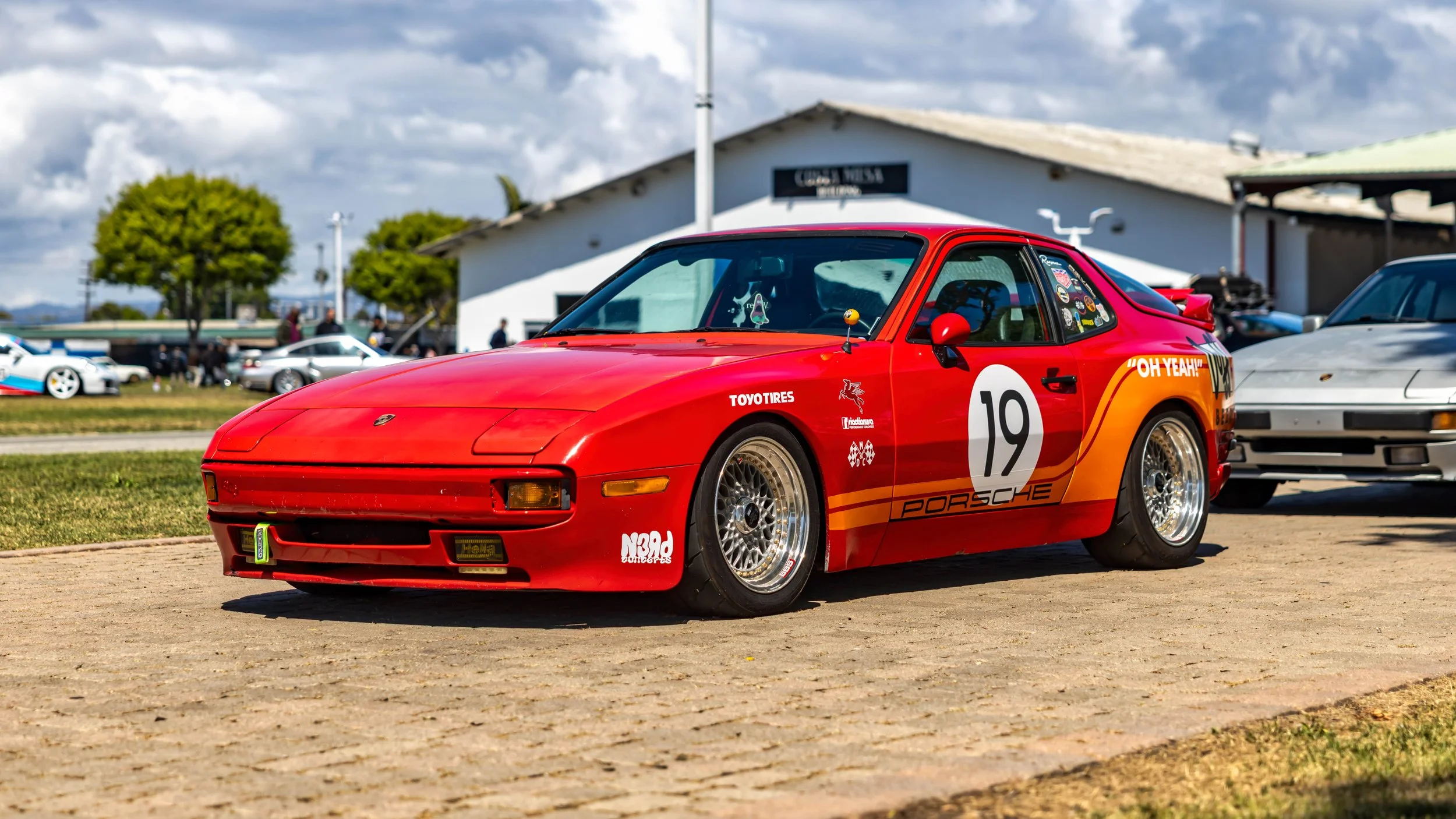 Air | Water 2025 — Orange County Fairgrounds — A red Porsche 944 with a vintage-style racing livery is parked on a paved lot at an outdoor car event. The car features a prominent number "19" roundel, "PORSCHE" lettering on the doors, a yellow-to-oran