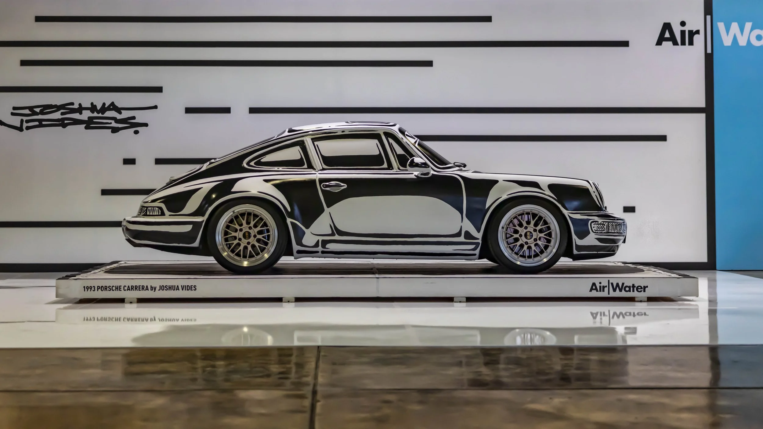 Air | Water 2025 — Orange County Fairgrounds — A side profile view of a 1993 Porsche 911 Carrera (964) art car by Joshua Vides, featuring a distinctive black-and-white, sketch-like livery. The car is presented on a white display platform at an 'Air |
