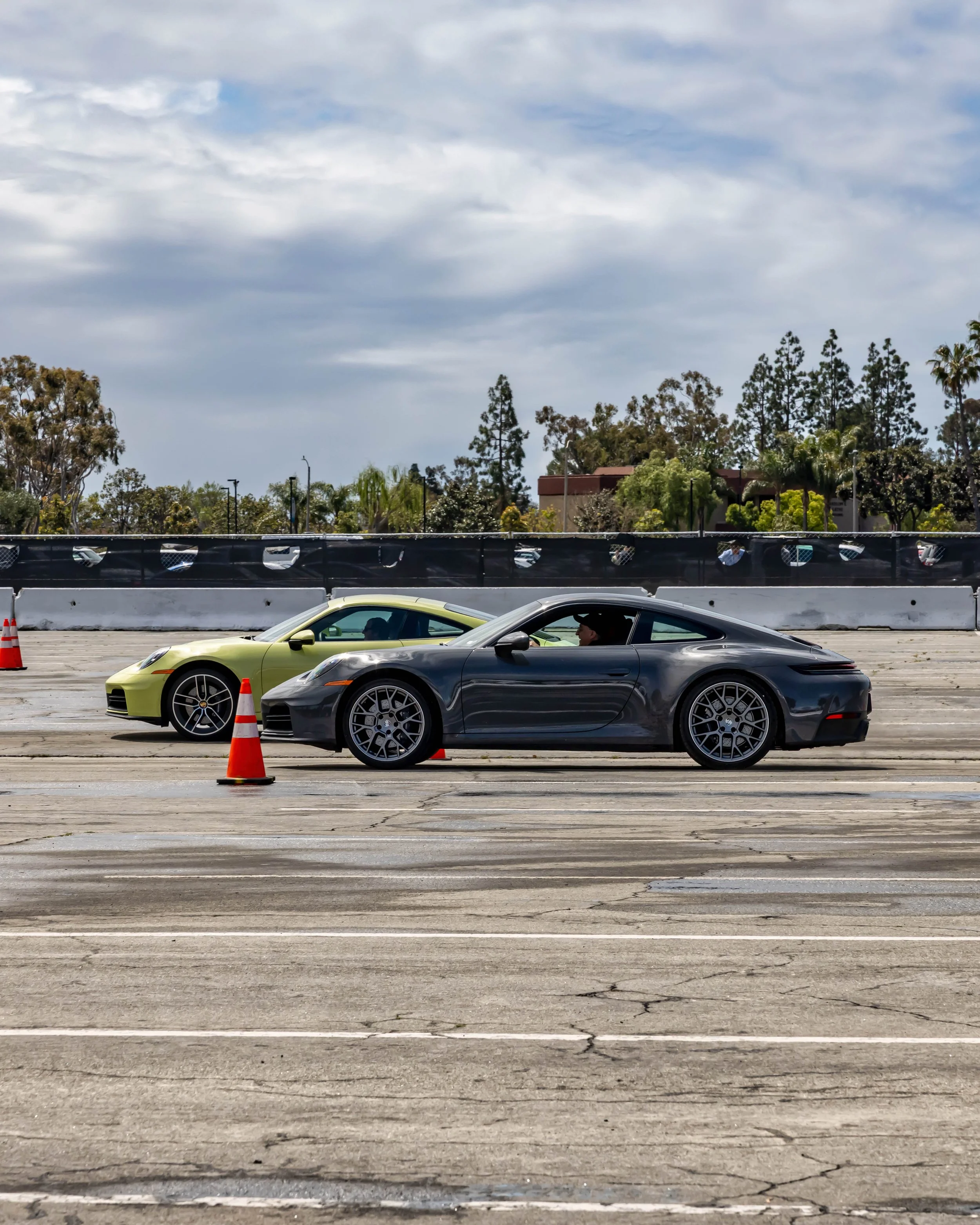 Air | Water 2025 — Orange County Fairgrounds — A profile view of a dark grey Porsche 911 Carrera (992) racing on a cracked asphalt lot next to a lime green 911. The scene is of a autocross course under an overcast sky. A driver is visible in the grey