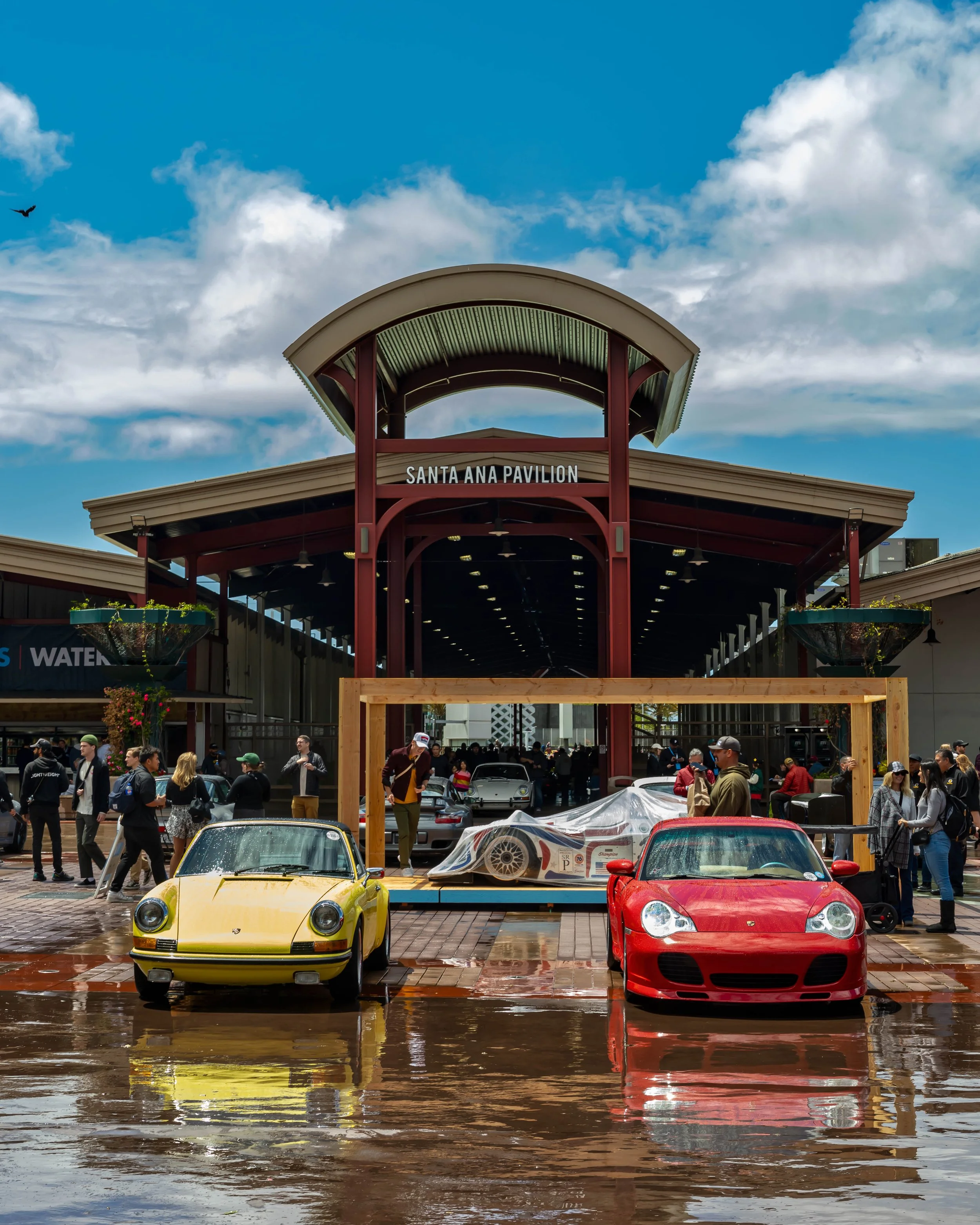 Air | Water 2025 — Orange County Fairgrounds — A red Porsche 911 GT3 (996) and a yellow classic Porsche 911 are parked in the foreground on wet brick pavement, reflecting their colors. Between them, a Champion Racing Porsche prototype is displayed on