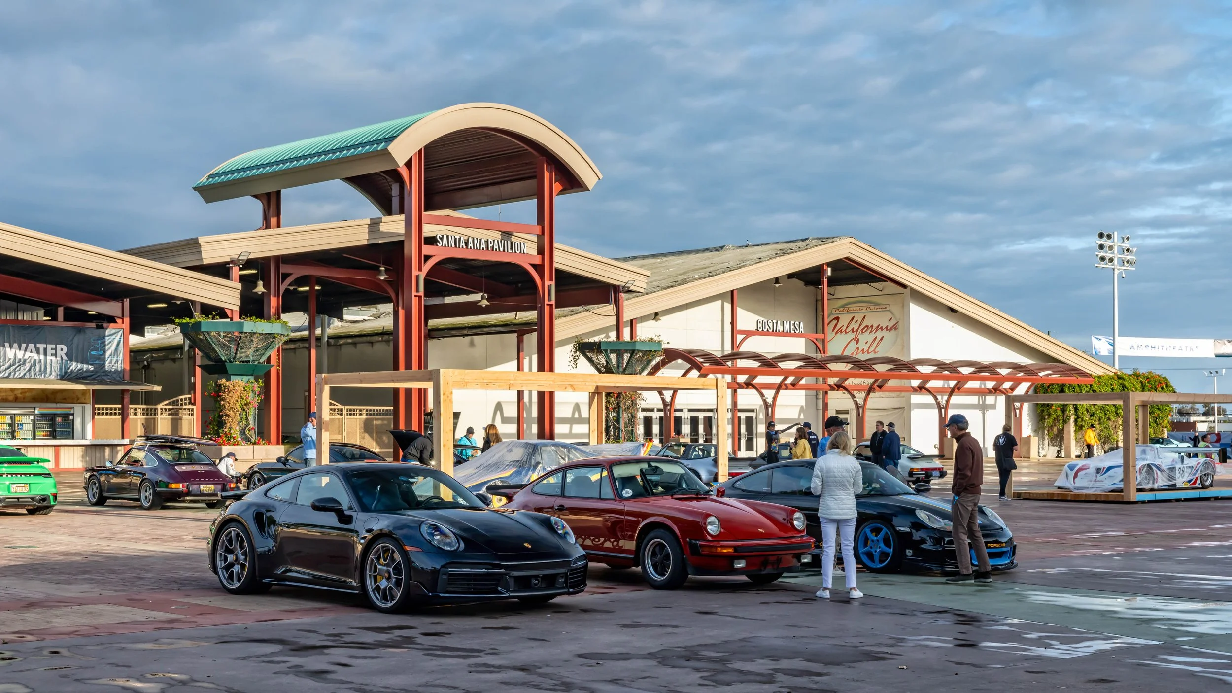 Air | Water 2025 — Orange County Fairgrounds — A gathering of various Porsche models, both classic and modern, are parked in an event space under an overcast sky. In the foreground, a black 992-generation Porsche 911 Turbo S is featured prominently, 