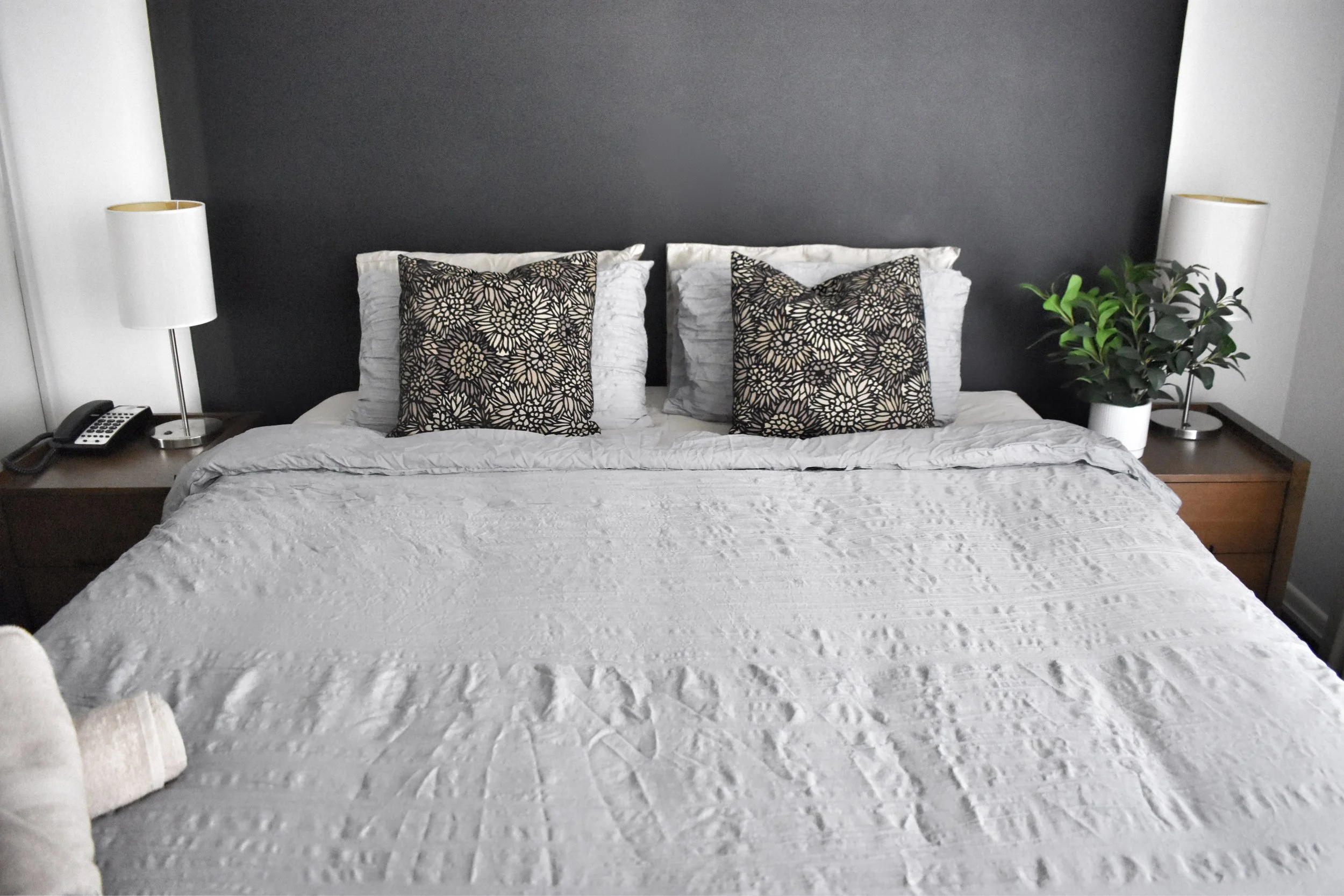 A neatly made bed with white textured bedding, two white pillows, two decorative pillows with black and beige floral patterns, flanked by two white bedside lamps on wooden nightstands, with a dark gray accent wall behind the bed and a green potted plant on the right side.