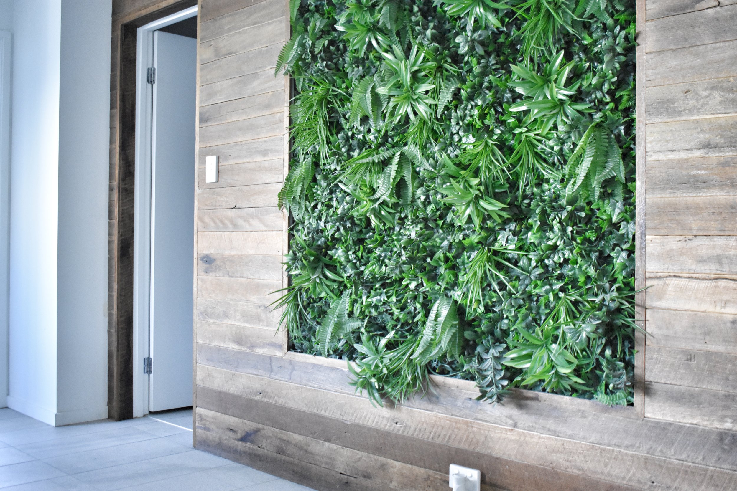 Indoor decorative wall with lush green plants in a wooden planter box next to a door and light-colored tiled floor.