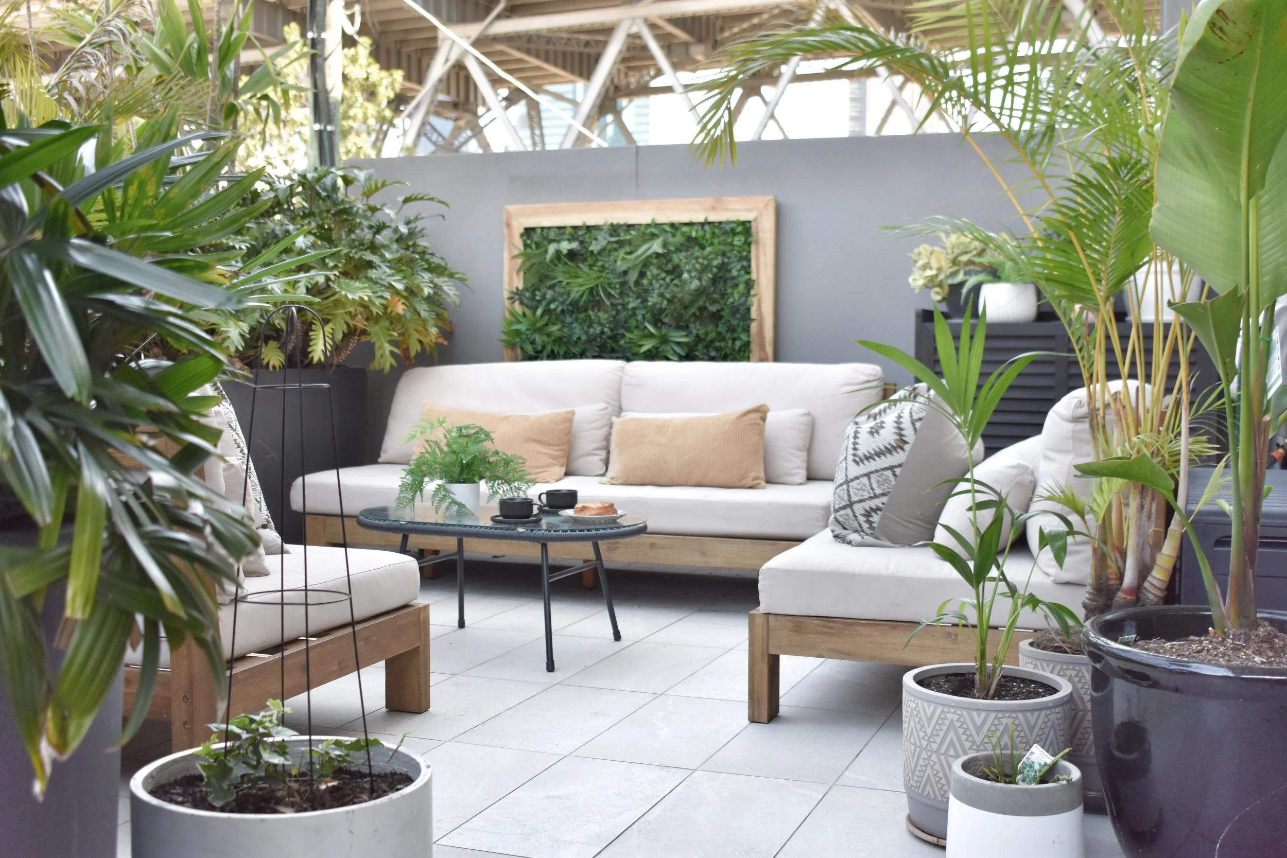 Indoor patio with white couches, wooden coffee table, and numerous potted plants, including large leafy tropical plants, against a gray wall with greenery art.