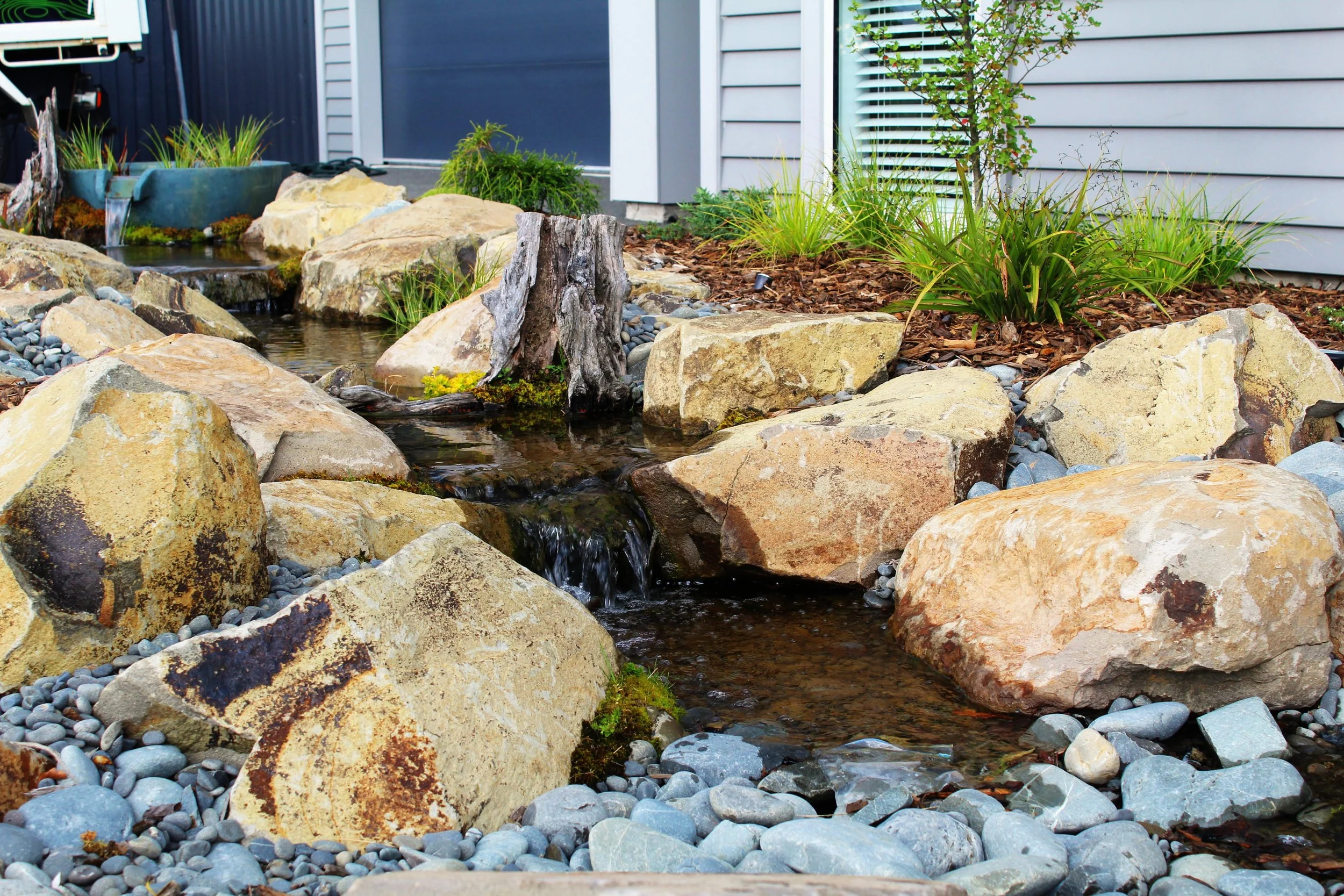 A landscaped garden feature with a small waterfall flowing over rocks, surrounded by large and small stones, with some green plants and mulch near the house.