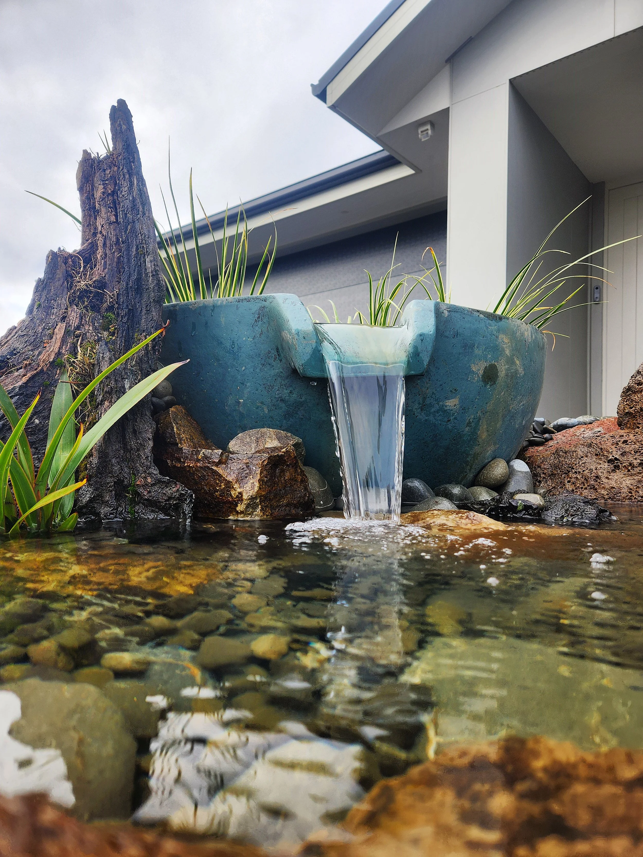 Close-up of a small artificial water fountain with water flowing into a shallow pond, surrounded by natural rocks and plants, with a house in the background.