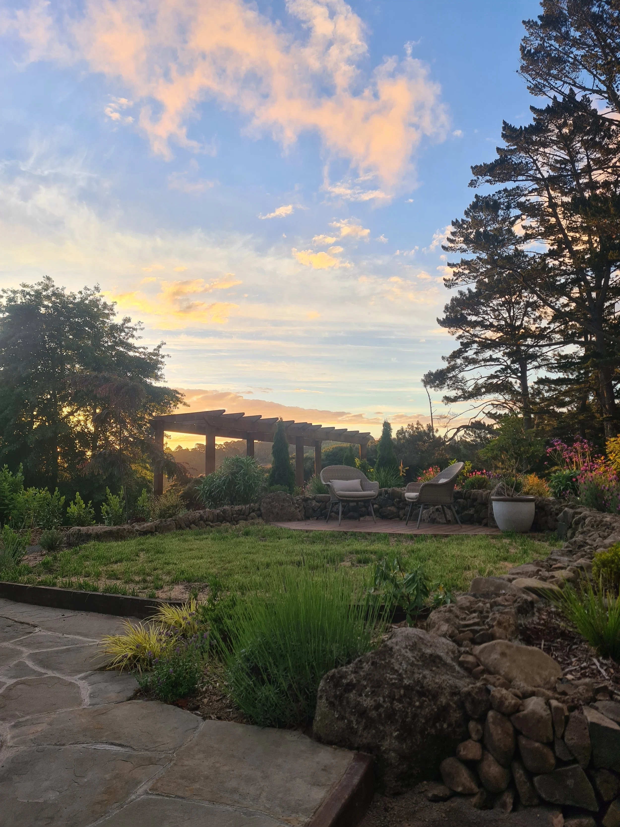 A peaceful backyard garden during sunset with a stone pathway, green grass, potted plants, and two chairs on a small patio, surrounded by trees and a wooden pergola.