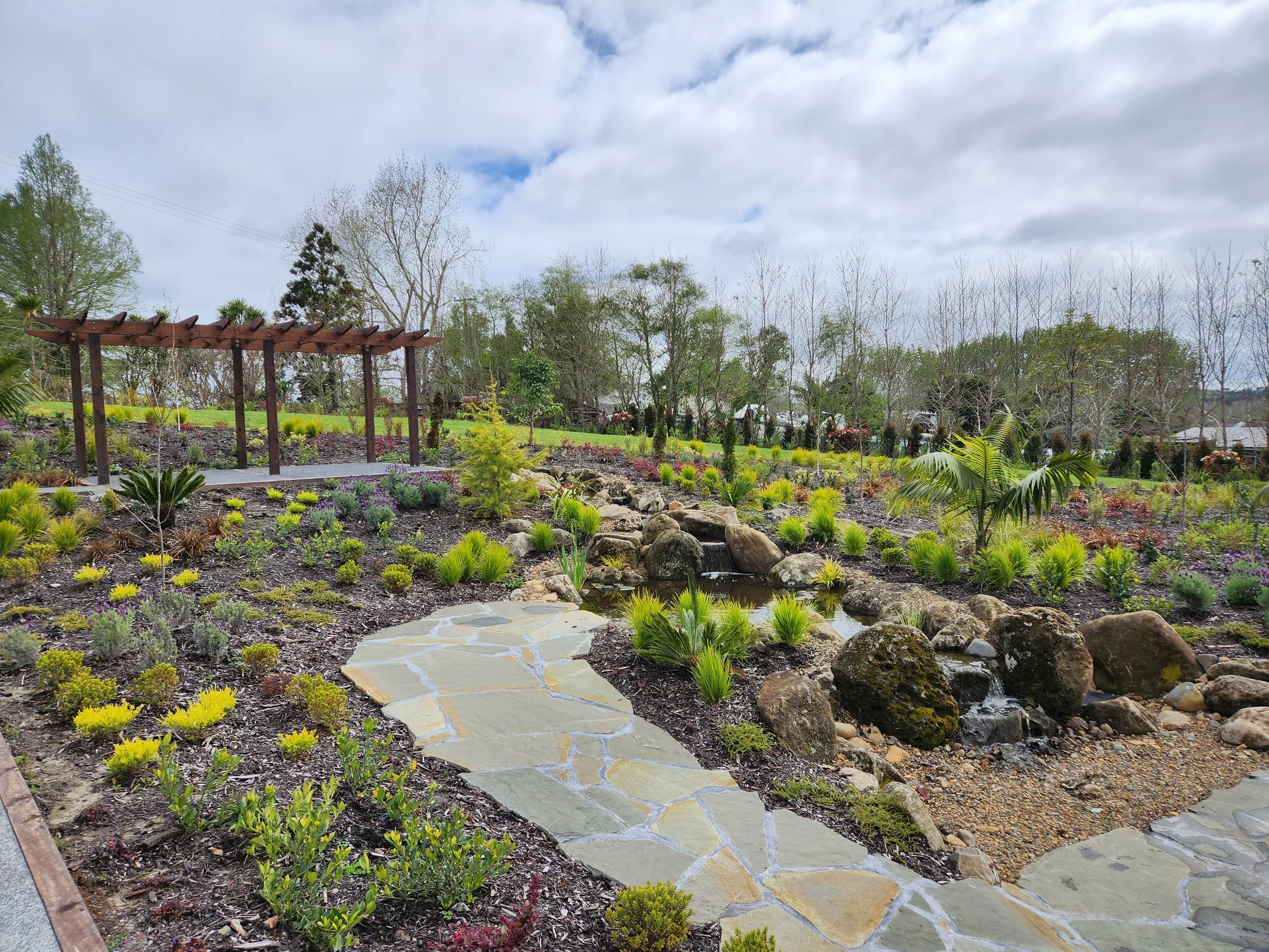 Beautiful landscaped garden with a stone pathway, small pond with rocks, tropical plants, and a wooden pergola under a cloudy sky.