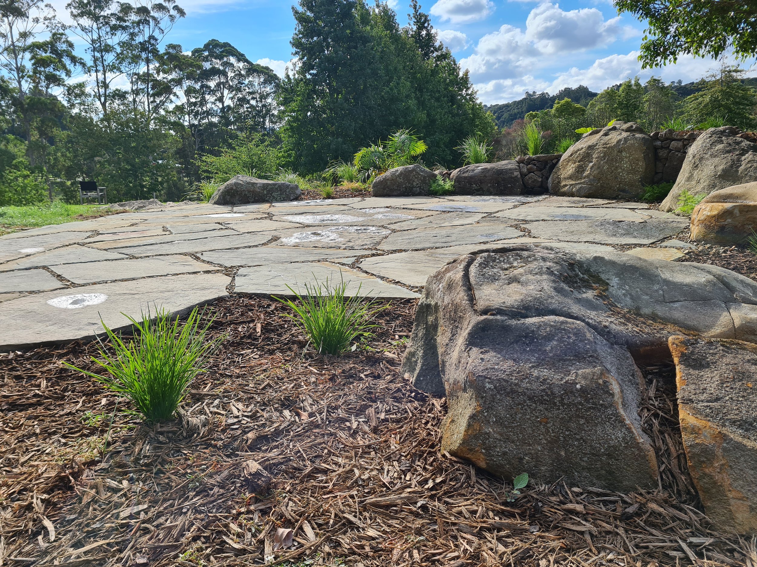 A stone pathway surrounded by large rocks and green plants, with trees in the background and a partly cloudy sky.