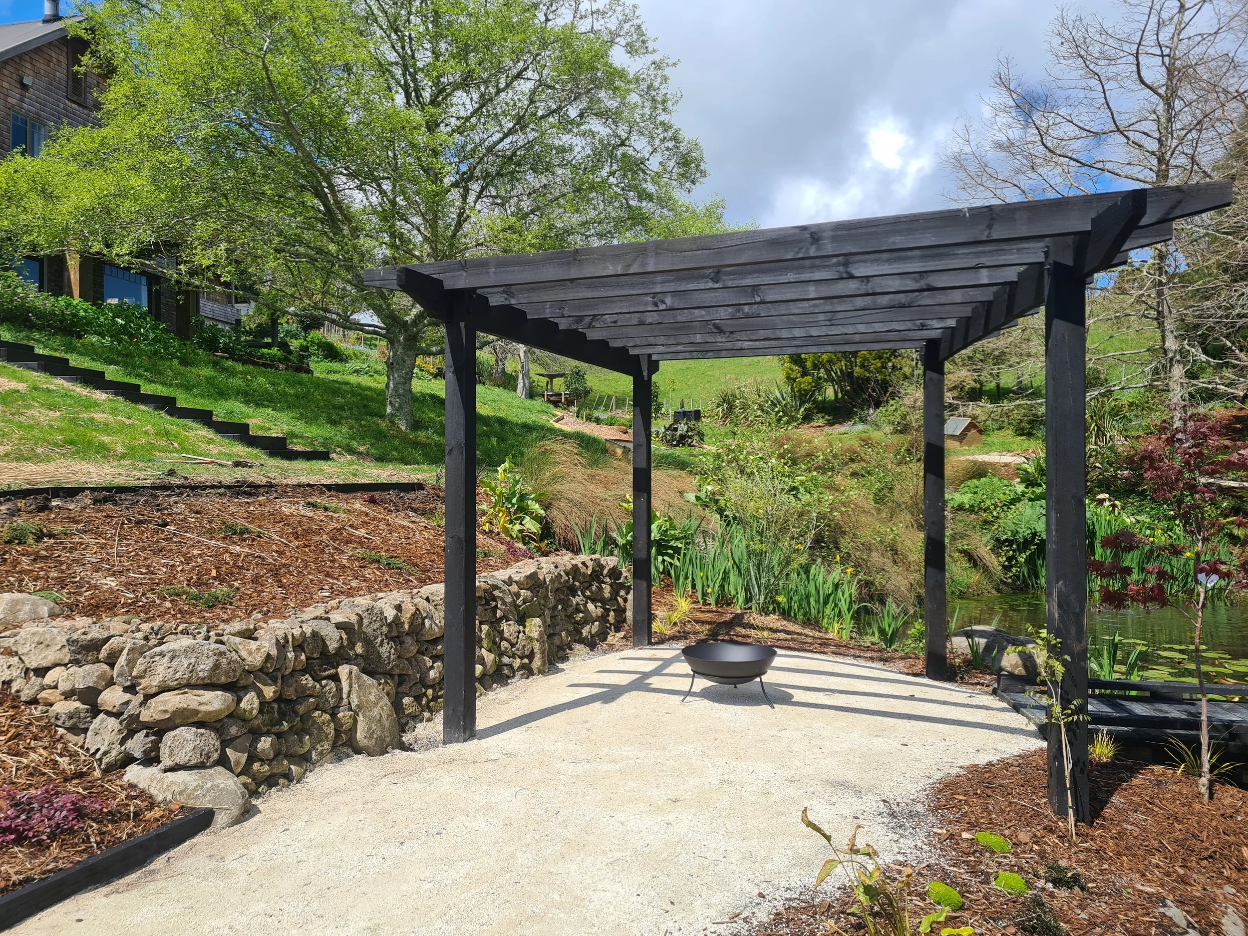 A black wooden pergola over a gravel patio in a lush garden with green plants, trees, and a rock wall.