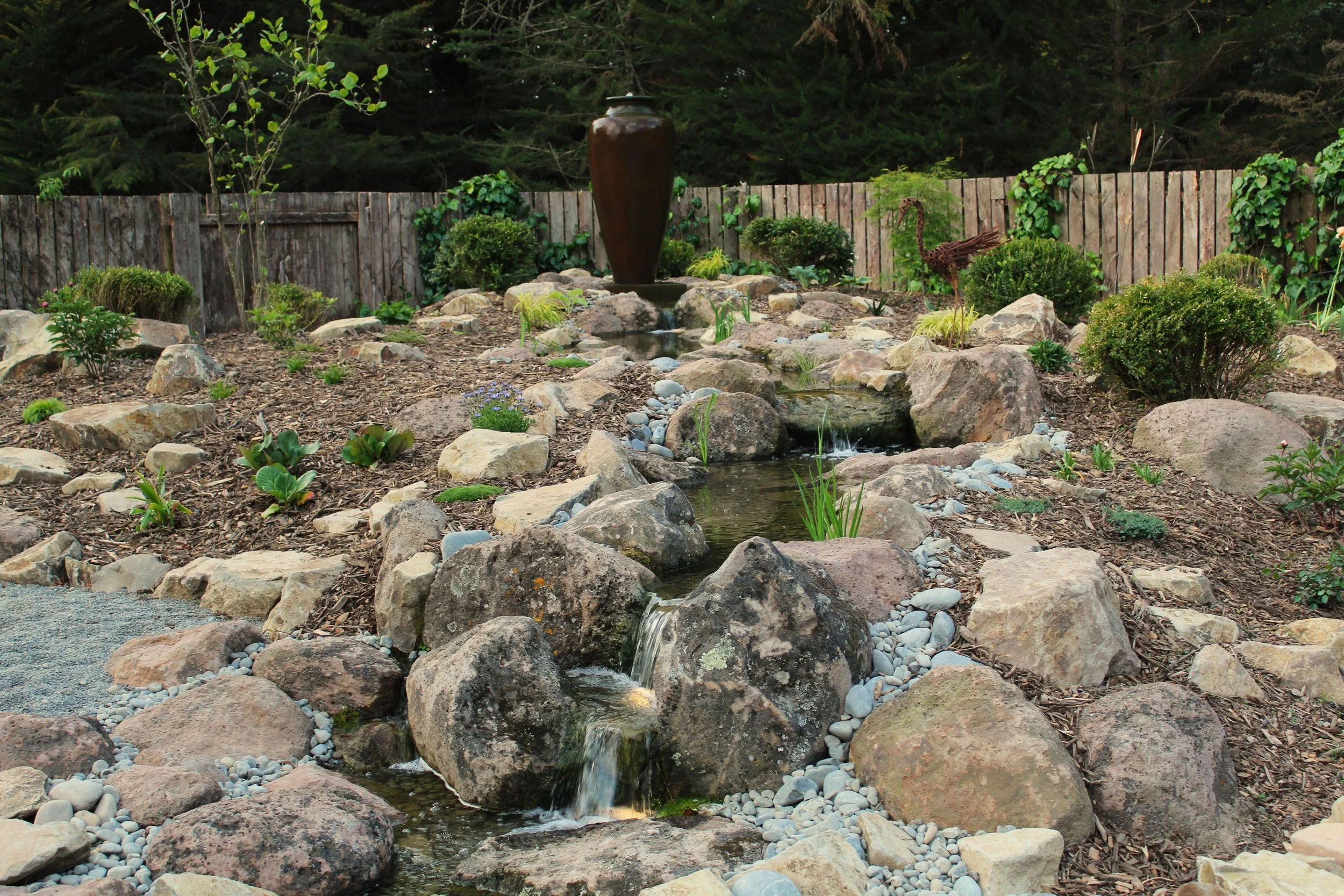 A landscaped backyard garden with a small, tiered rock garden and a water feature, including a tall brown vase fountain, surrounded by various plants, shrubs, and a wooden fence in the background.