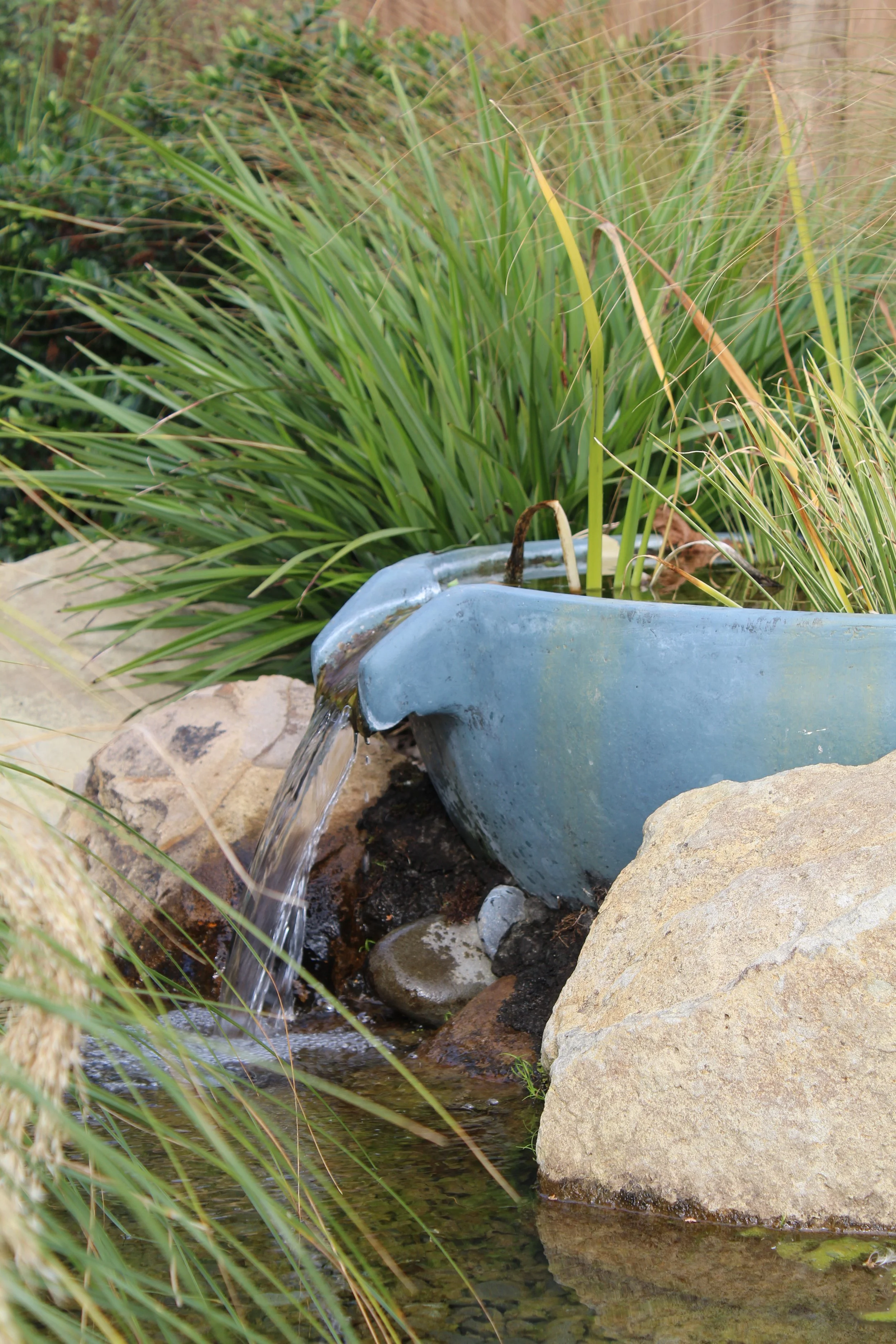 Pondless waterfall spilling from a ceramic urn into a rock-edged garden pond surrounded by tussock grasses