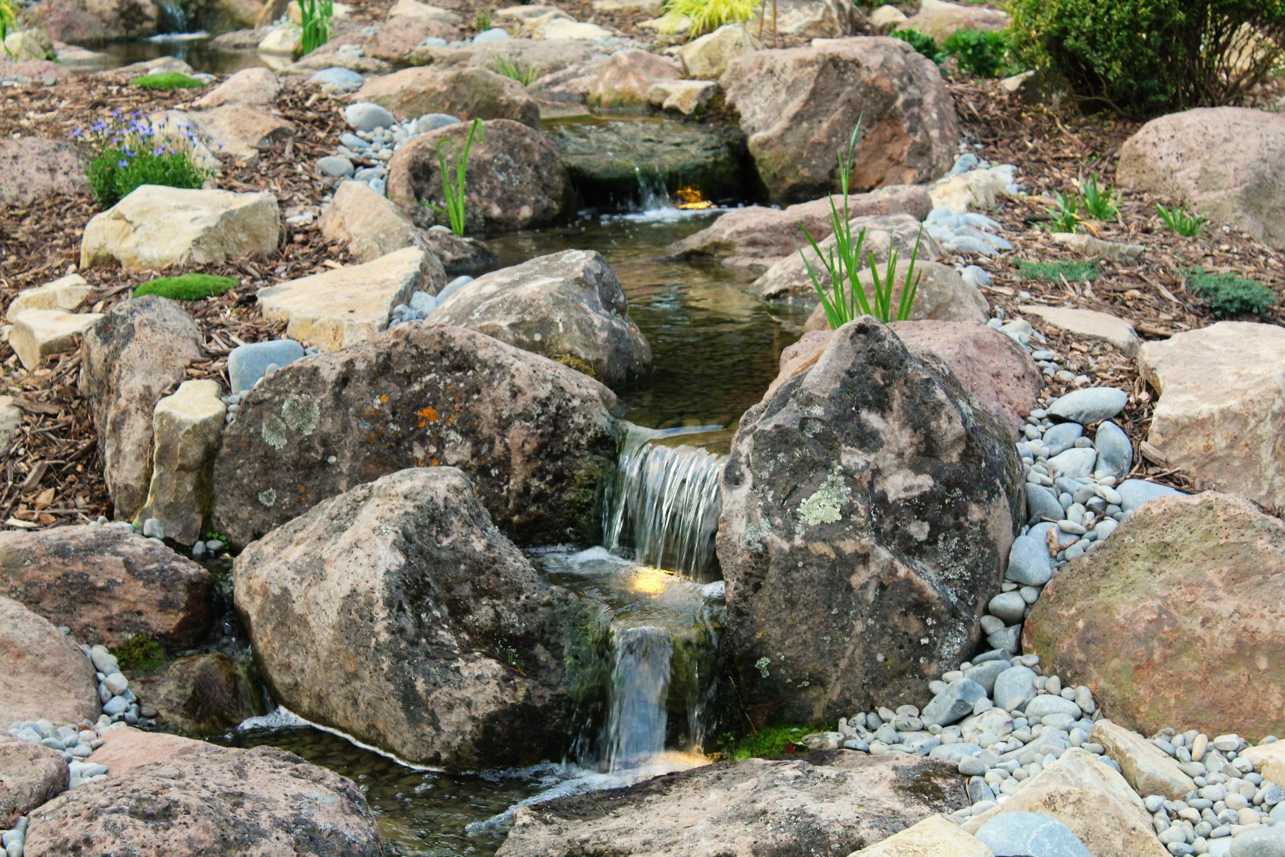 A small, artificial stream with cascading water surrounded by various rocks and pebbles in a landscaped garden.