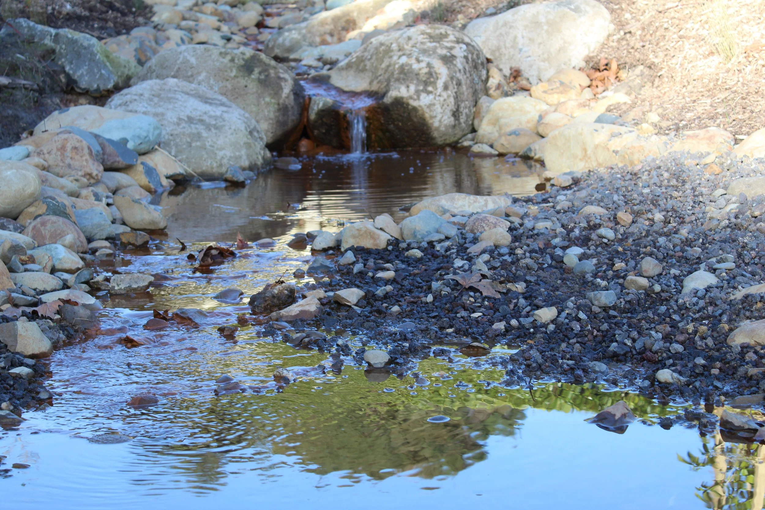 A small stream flowing over rocks and pebbles in a natural outdoor setting.