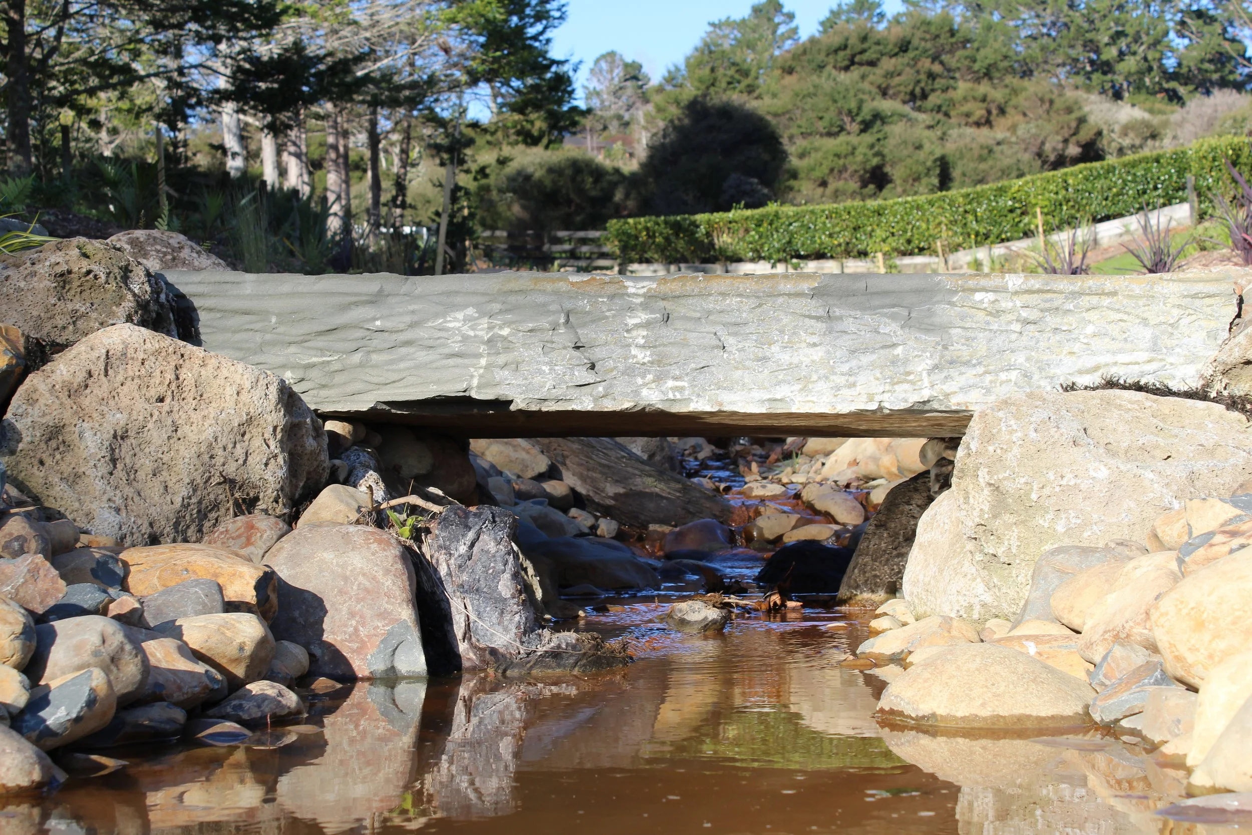 A small stream flowing under a flat stone bridge with large rocks on either side. In the background, there are trees, shrubs, and a clear blue sky.
