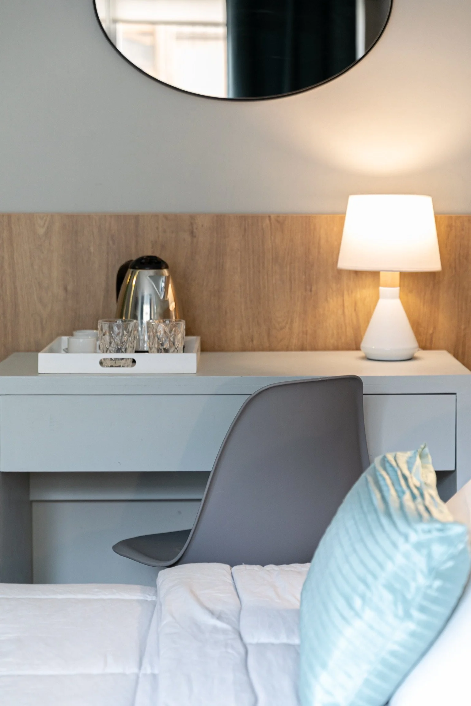 Habitación con una mesa, espejo redondo, lámpara encendida, hervidor de agua y varias tazas. Una silla gris y una cama con cojines azules.