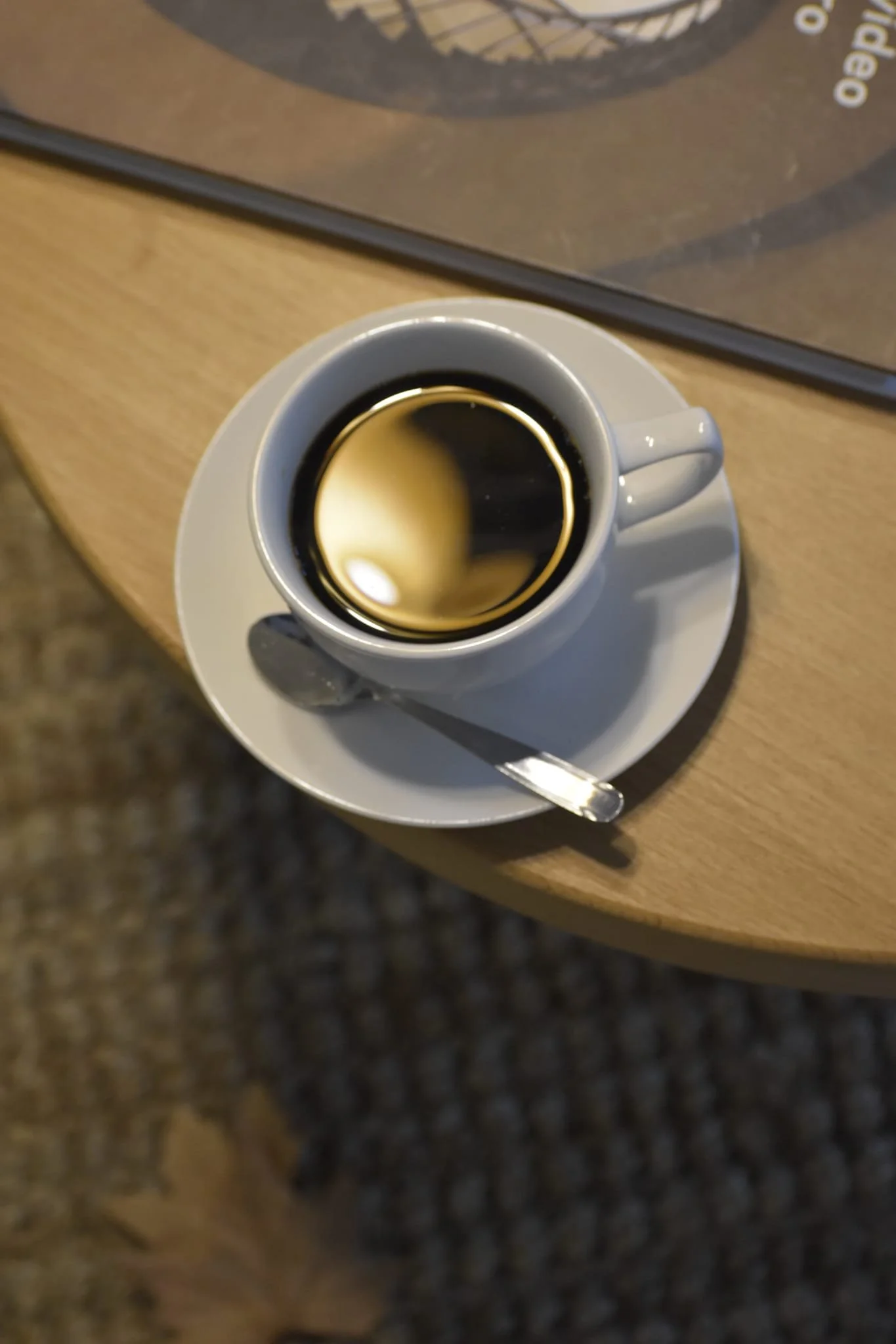 Taza de café negro sobre un plato blanco con una cuchara sobre una mesa de madera clara.