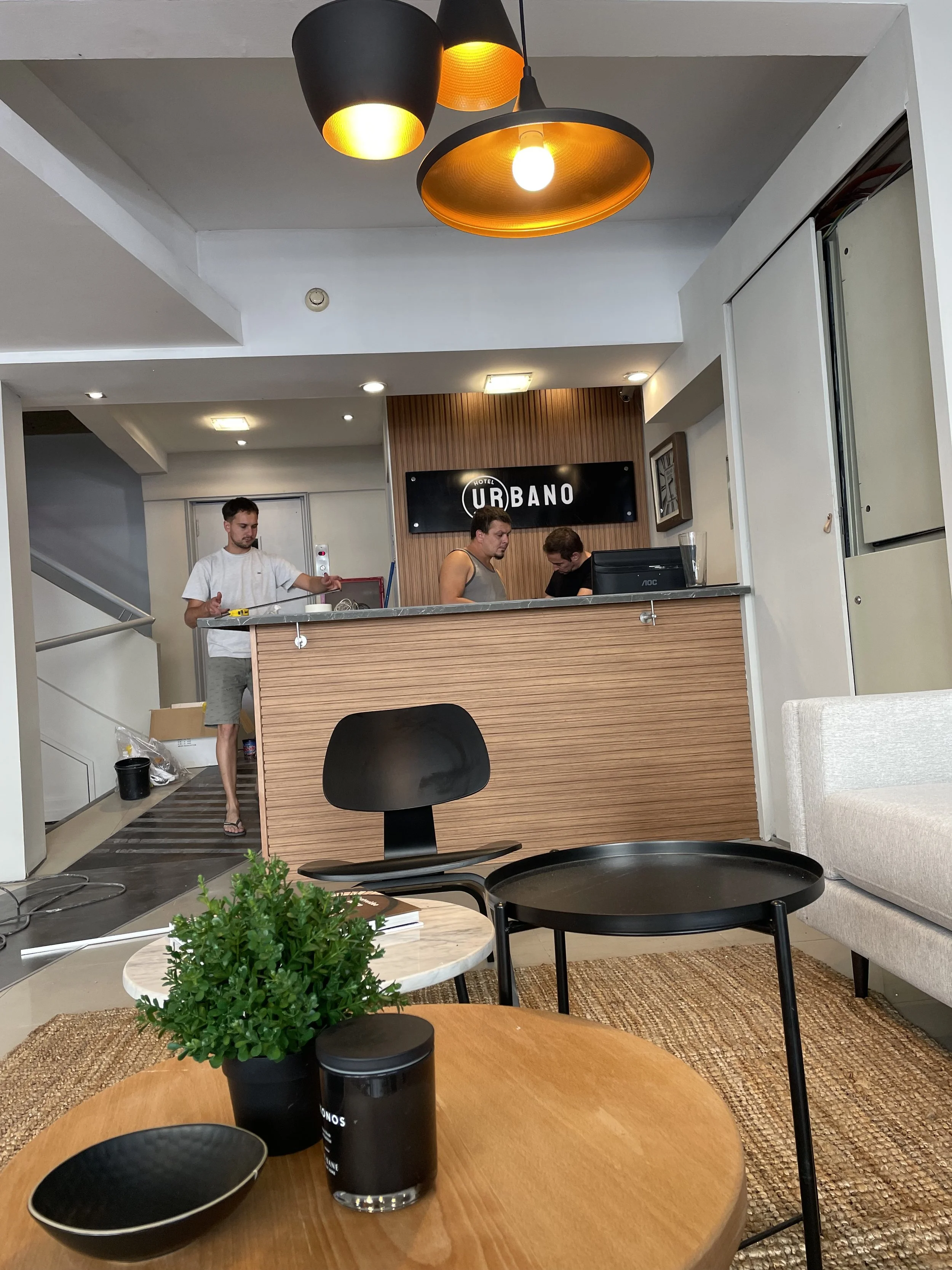 Lobby de un hotel con personas en la recepción y decoración moderna, incluyendo una planta y mesa en el primer plano. Lámparas modernas cuelgan del techo.