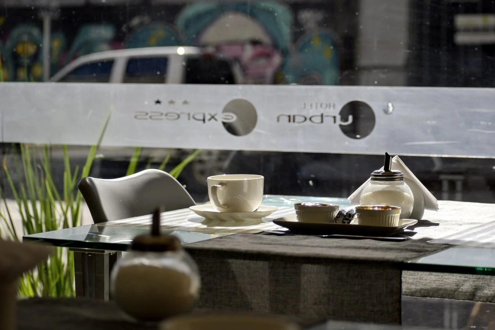 Mesa de cafetería con taza de café, azucarero, y vista a la calle a través de una ventana con el texto 'Hotel Urban Express'.