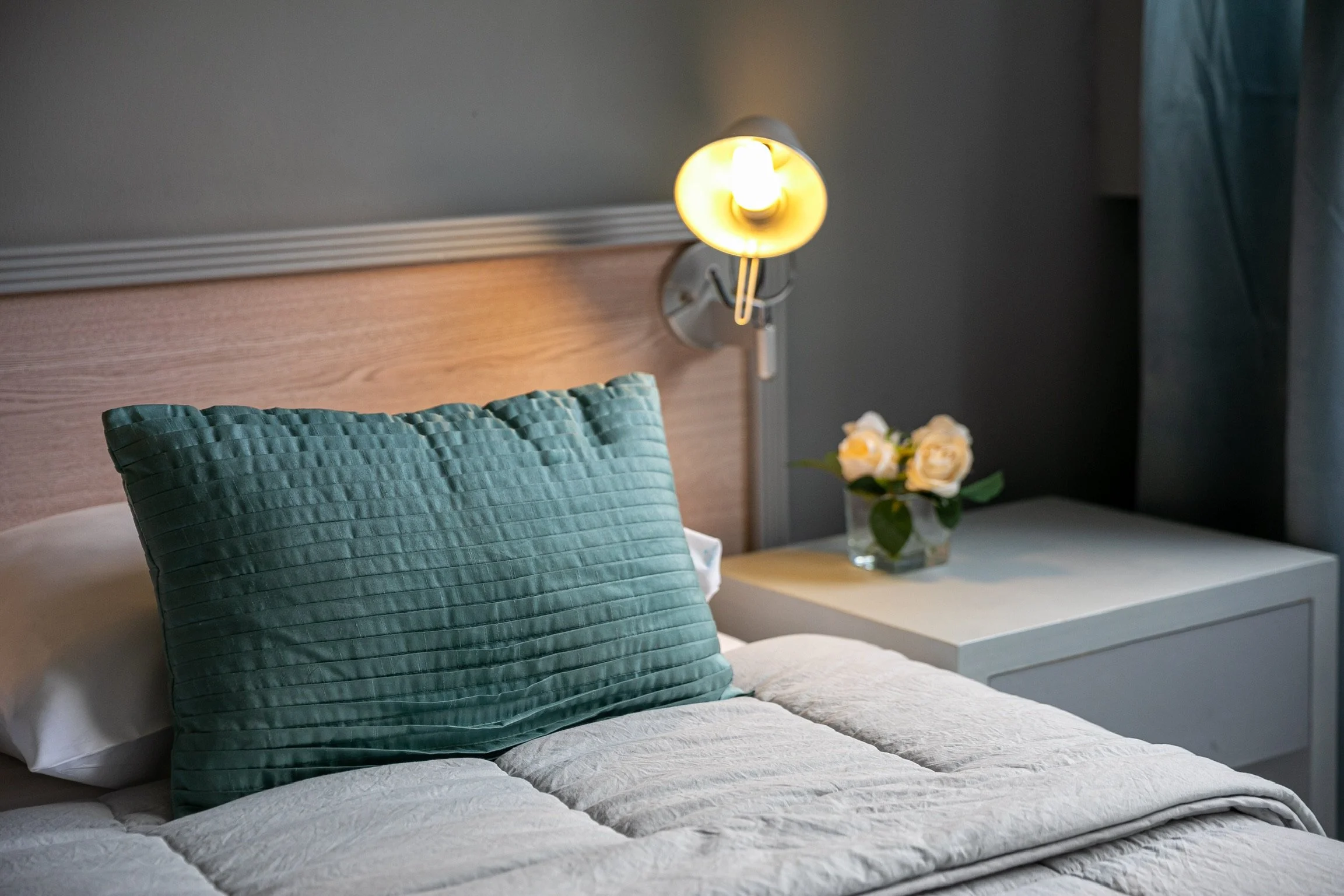 Habitación con cama y cojín verde, lámpara de pared encendida y mesa de noche con rosas blancas.