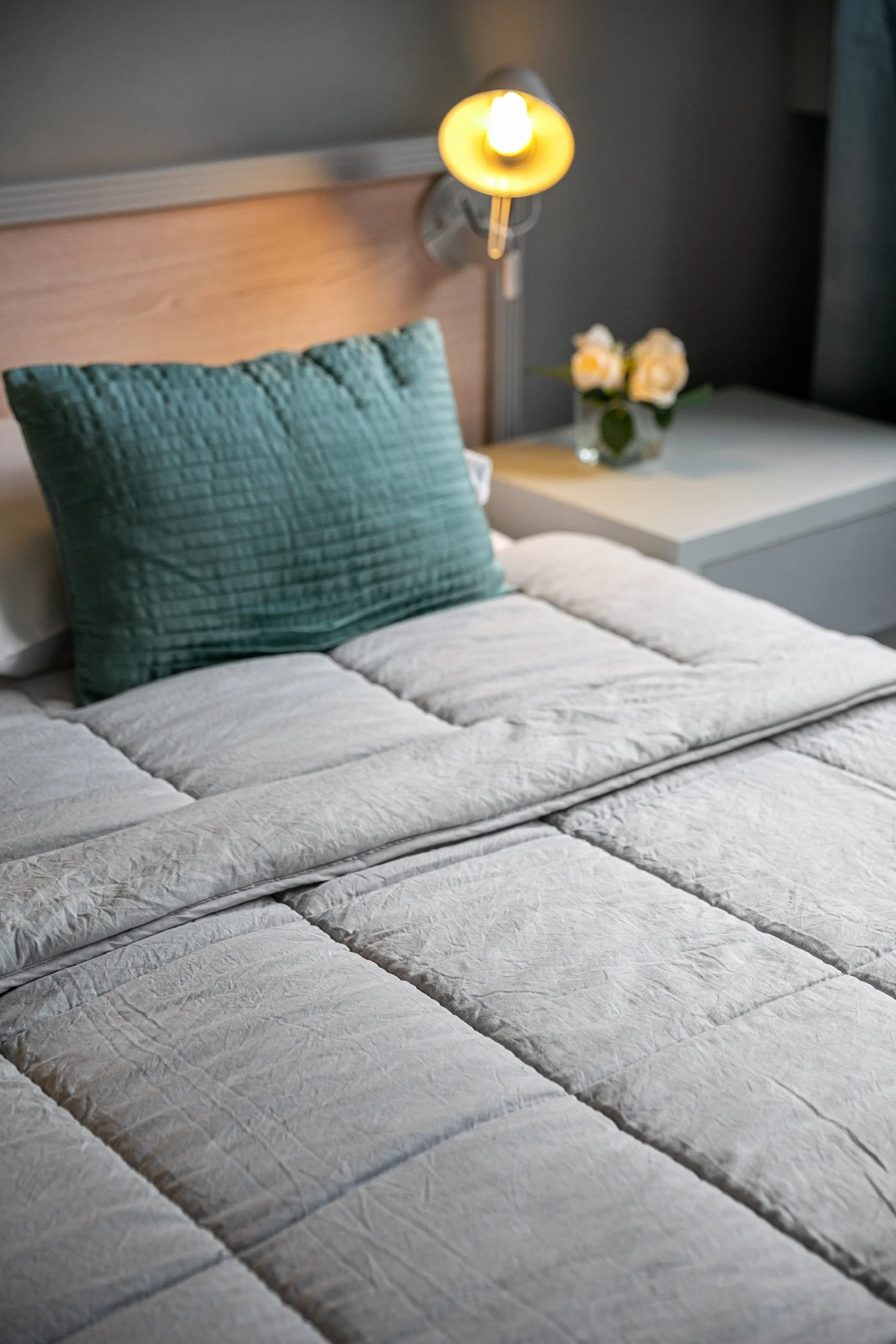 Cama con edredón gris, almohada verde, lámpara de pared encendida, y mesa de noche con flores blancas en un jarrón.