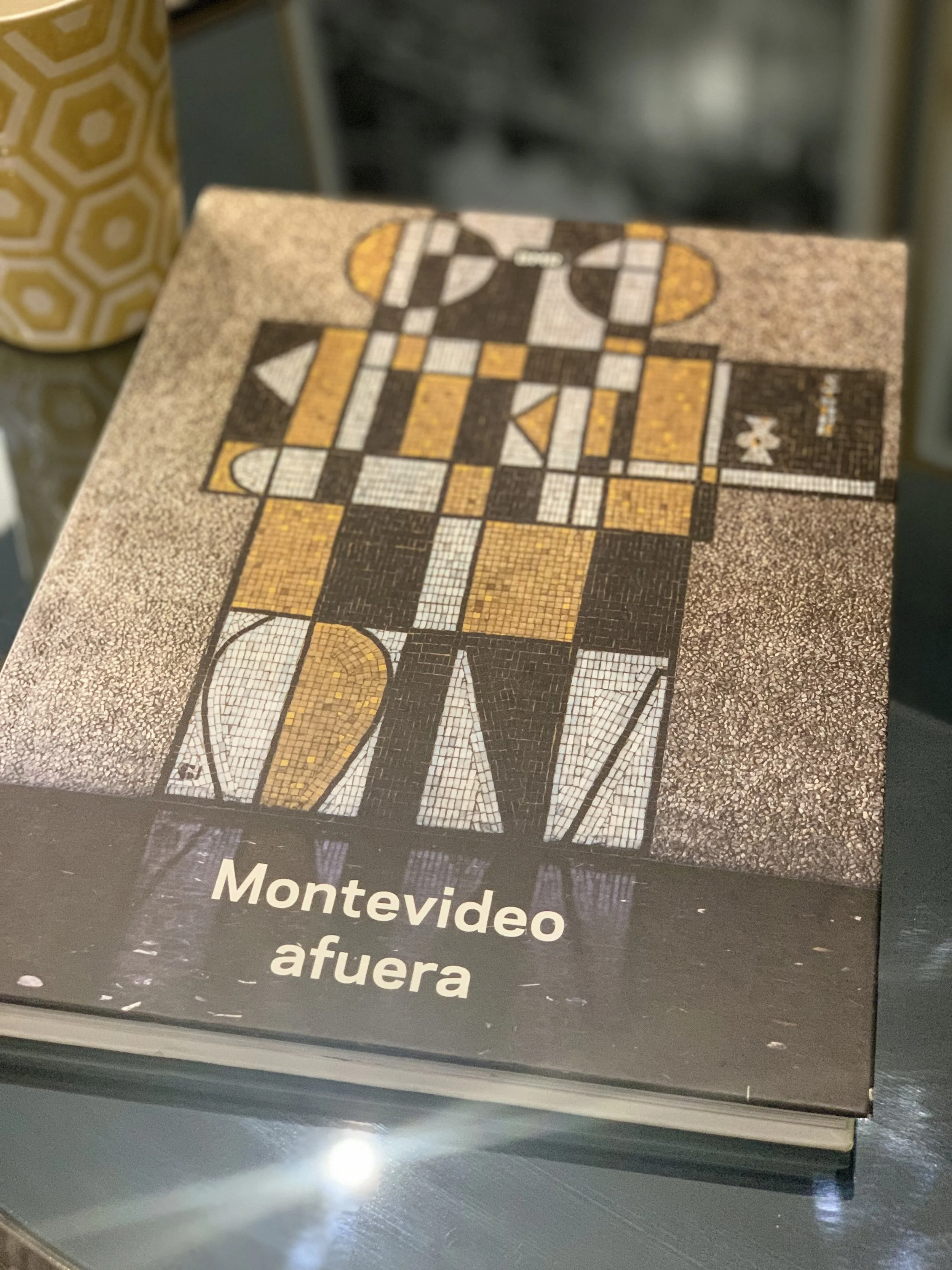 Libro titulado "Montevideo afuera" con portada que muestra un diseño artístico geométrico en blanco, negro y amarillo. Fondo oscuro. Taza decorativa a la izquierda.