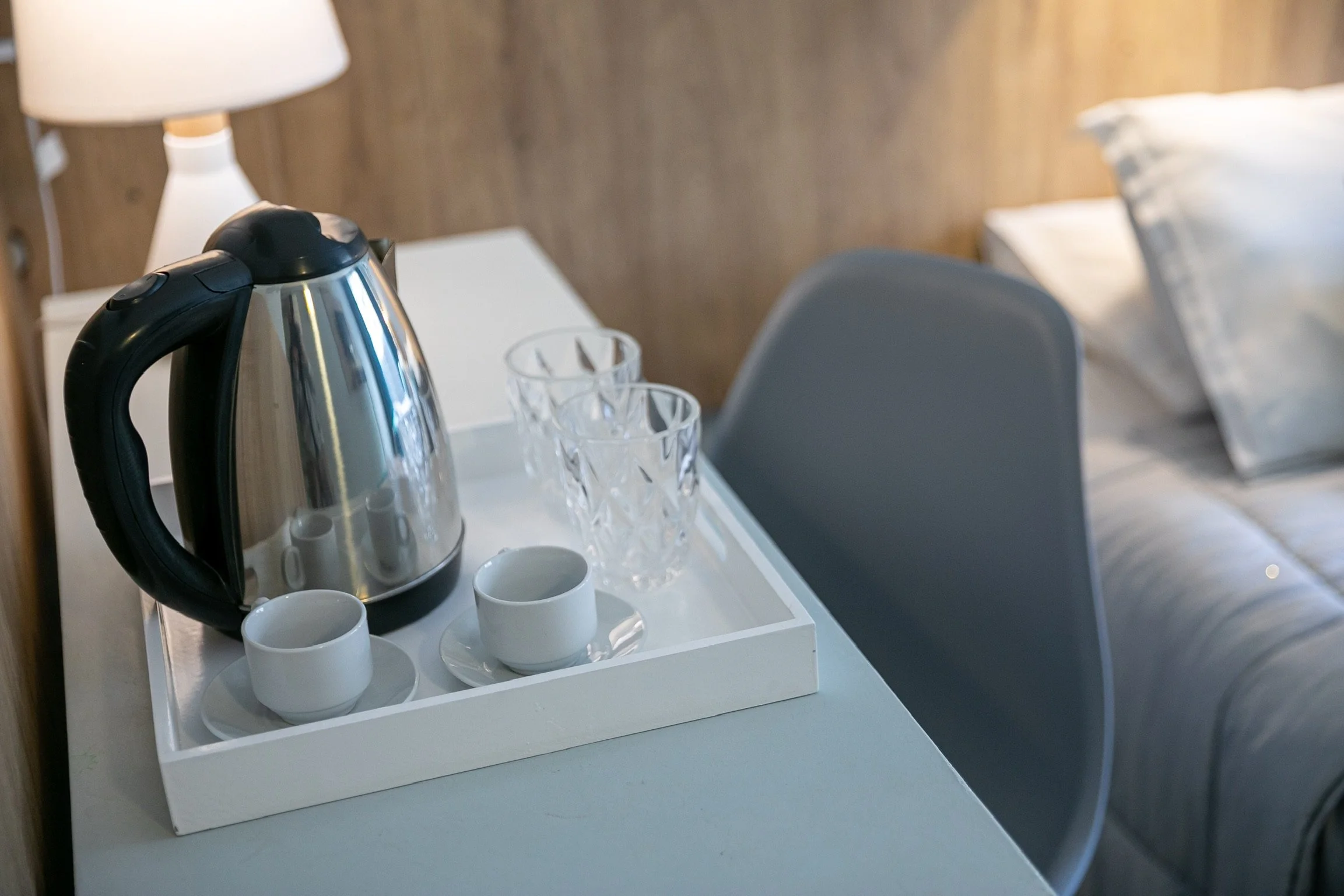 Tetera eléctrica de acero inoxidable en una bandeja blanca con tazas y vasos, junto a una lámpara y una cama en una habitación simple.