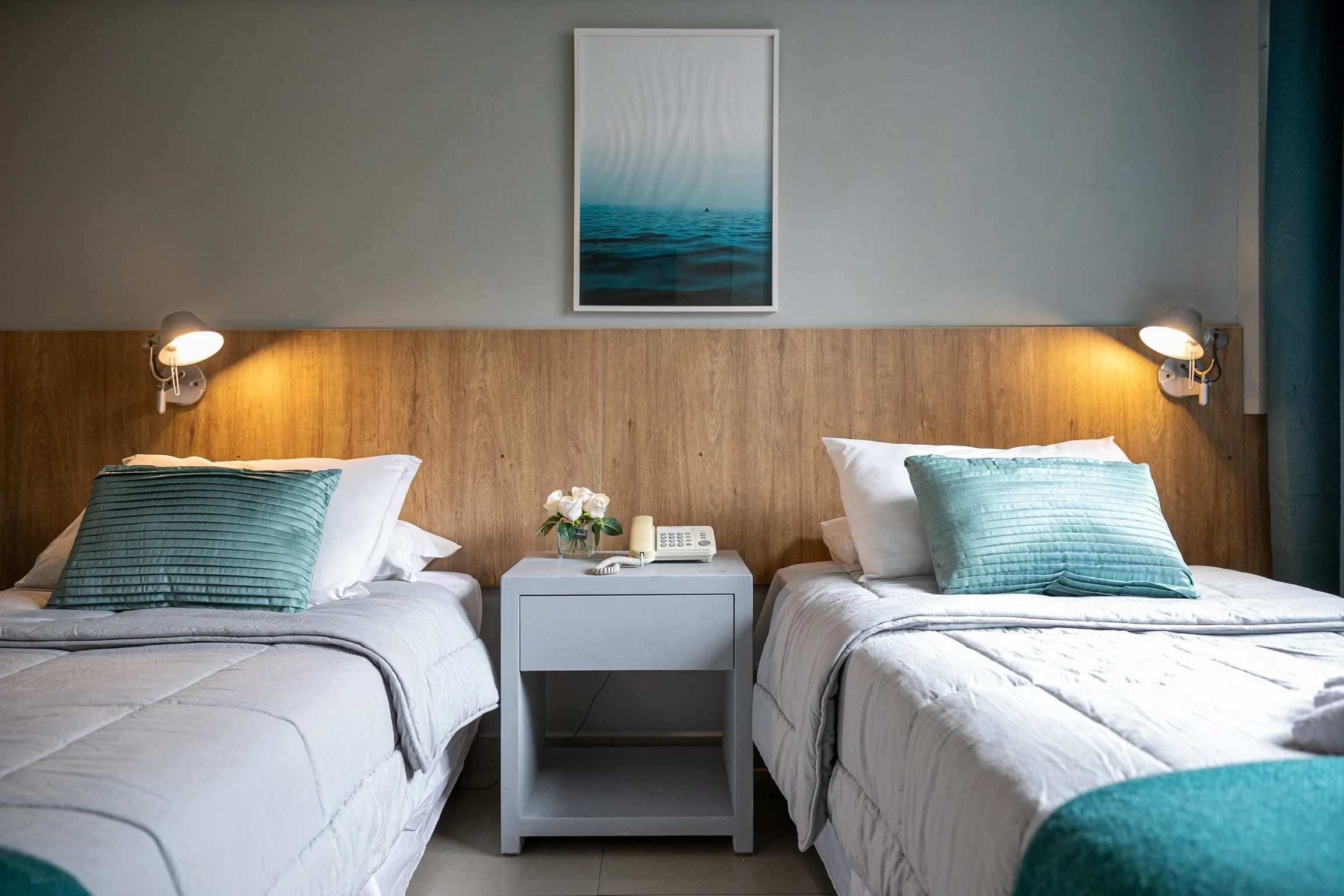 Habitación moderna con dos camas individuales con almohadas turquesas, una mesita de noche gris con teléfono y flores, lámparas de pared, y cuadro de paisaje marino.