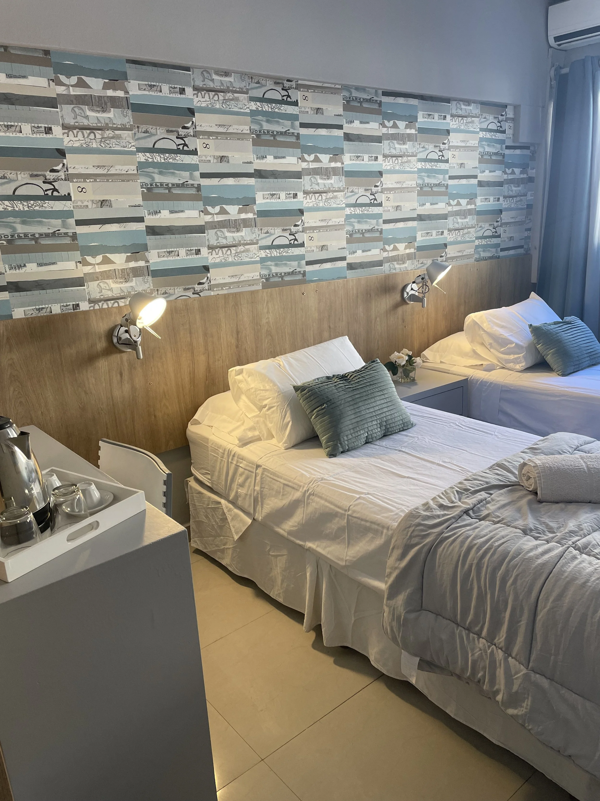 Habitación de hotel con dos camas individuales, ropa de cama blanca y cojines azul claro. Las paredes tienen papel tapiz de diseño moderno. Hay una mesita de noche con lámpara y una bandeja con tazas y hervidor de agua sobre un mueble gris. Aire acon