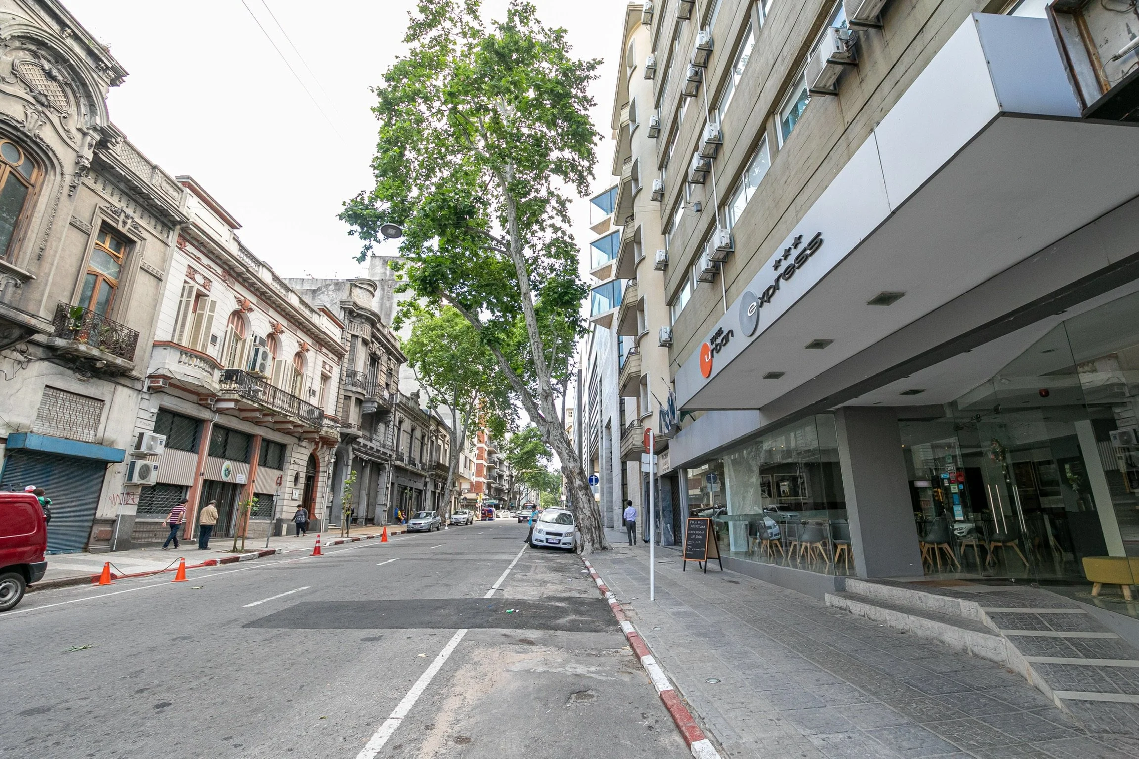 Vista de una calle urbana con edificios antiguos y modernos, árboles y algunos peatones. Hay un automóvil estacionado y conos naranjas a lo largo de la calle. Un negocio con ventanales ocupa la planta baja de uno de los edificios.