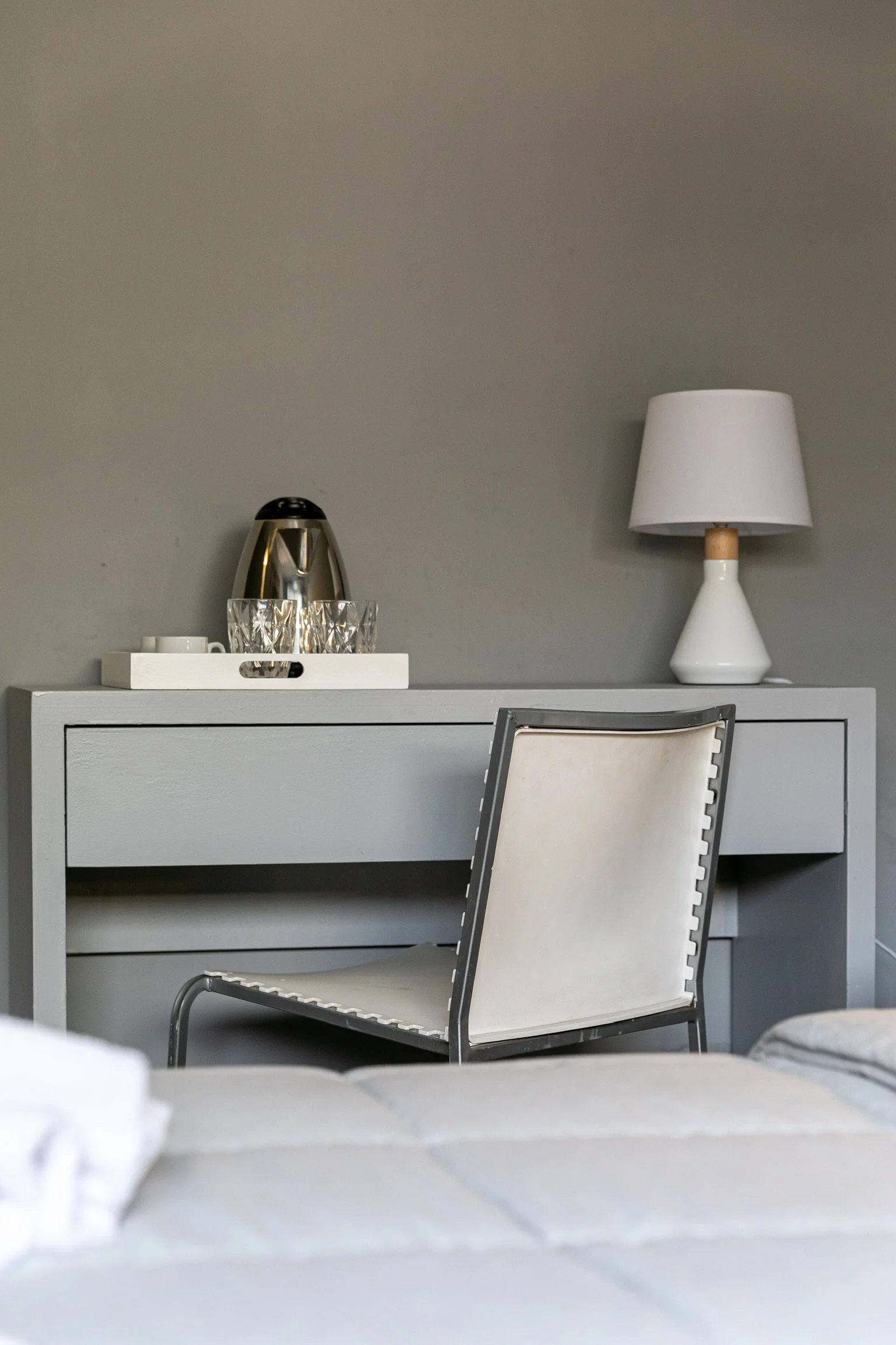 Mesa gris con silla blanca, lámpara de mesa, bandeja con tazas y tetera en habitación.