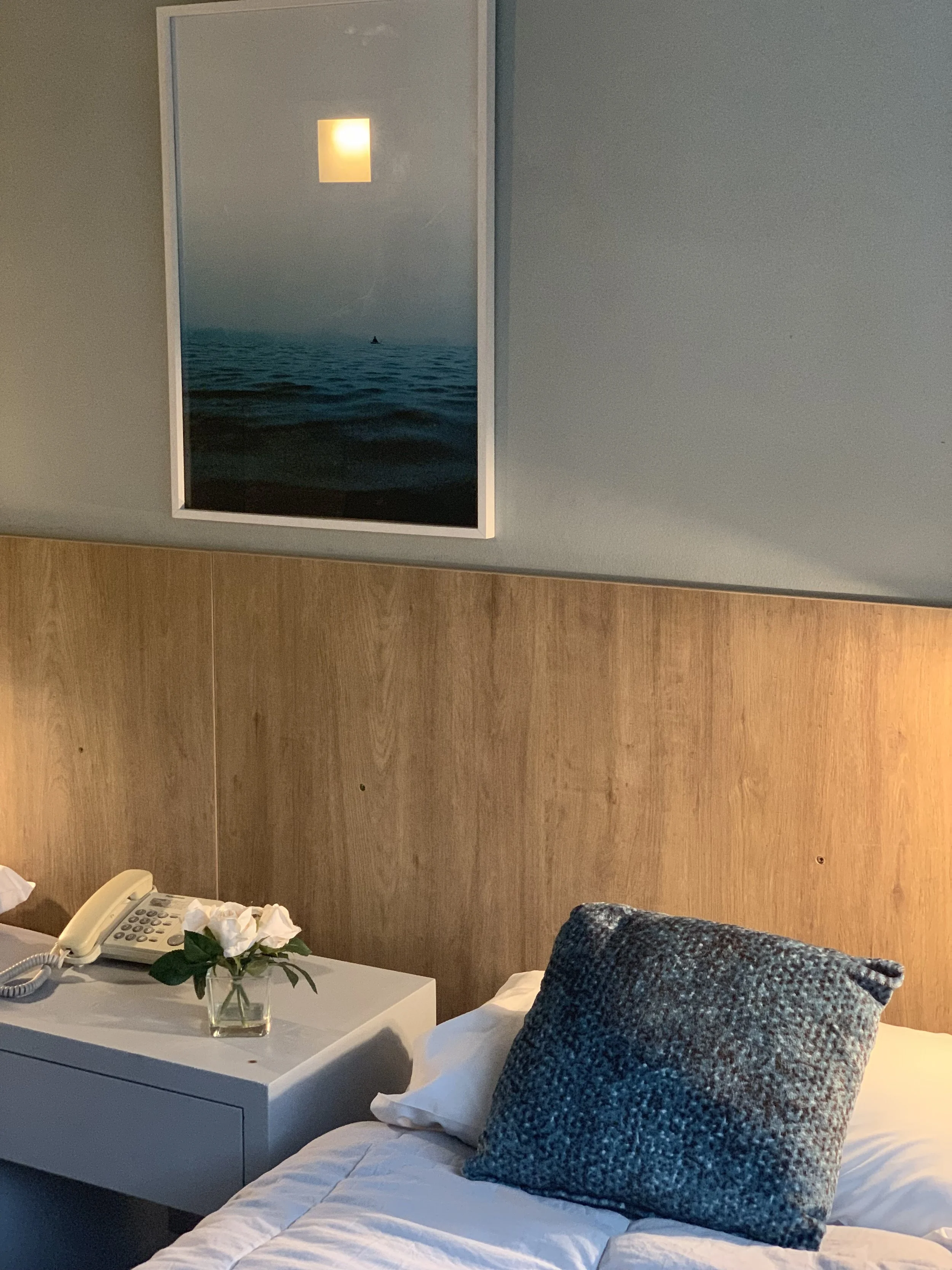 Habitación con cama, cojín azul, mesita con teléfono, flores en un jarrón y cuadro de paisaje marino en la pared.